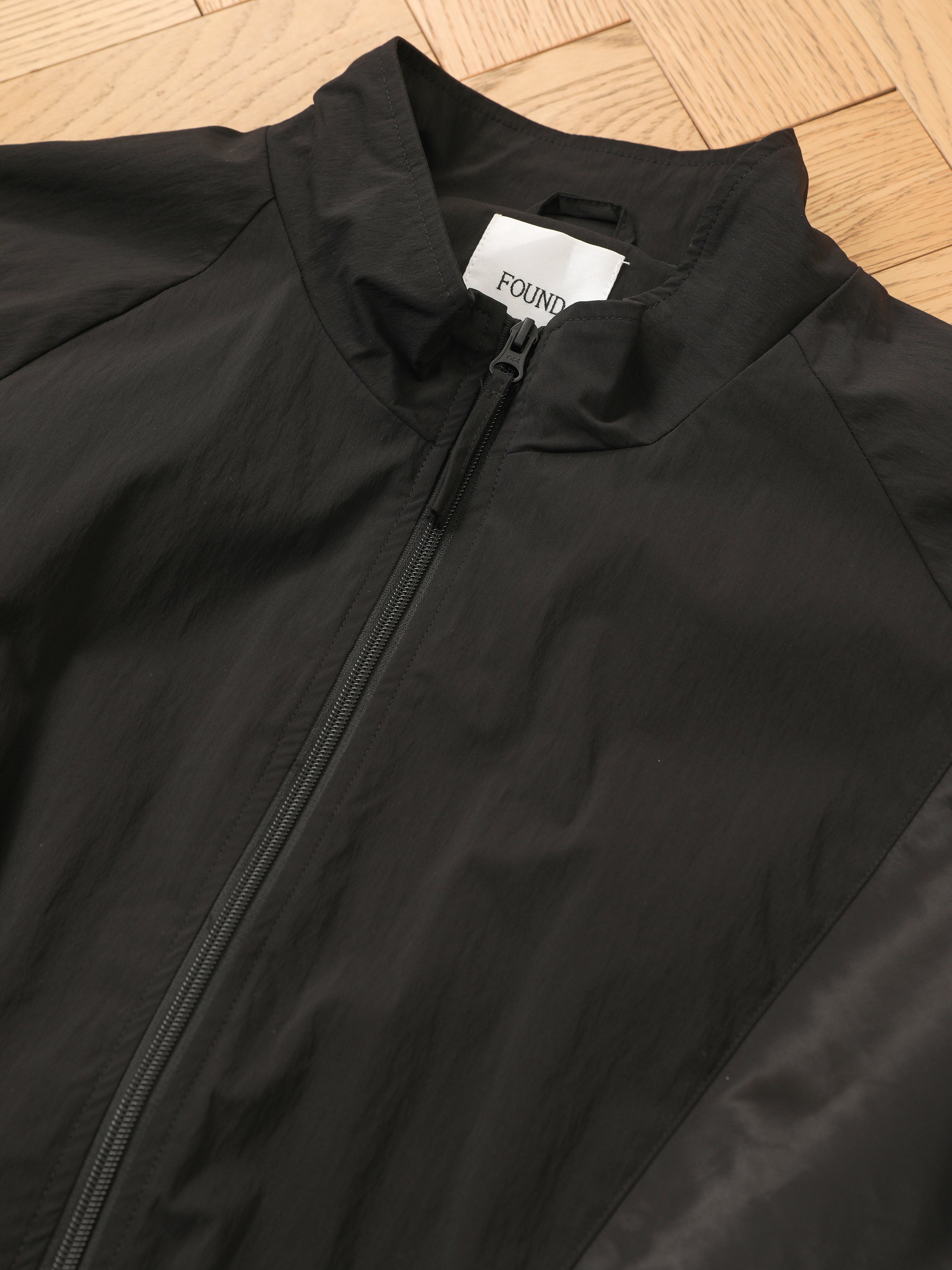 The Pine and Rose Jacket by FOUND is a black unisex windbreaker with a standing collar, displayed flat on a wooden floor. The inner label shows the brand name "FOUND.