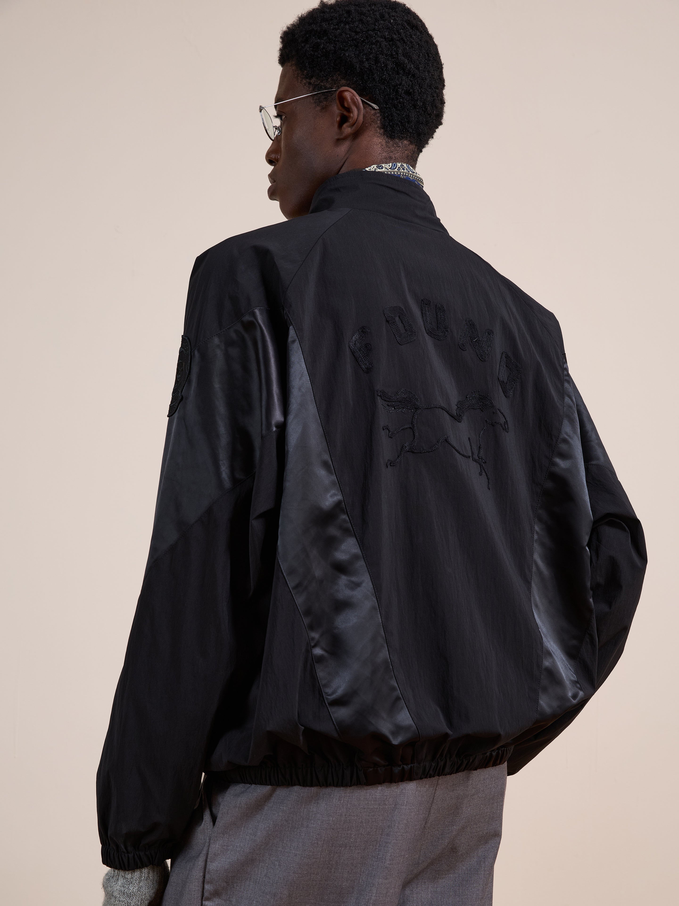 A person wears the FOUND Pine and Rose Jacket, a black vintage sportswear-inspired track jacket with satin panels and an embroidered horse design on the back, standing against a plain background.
