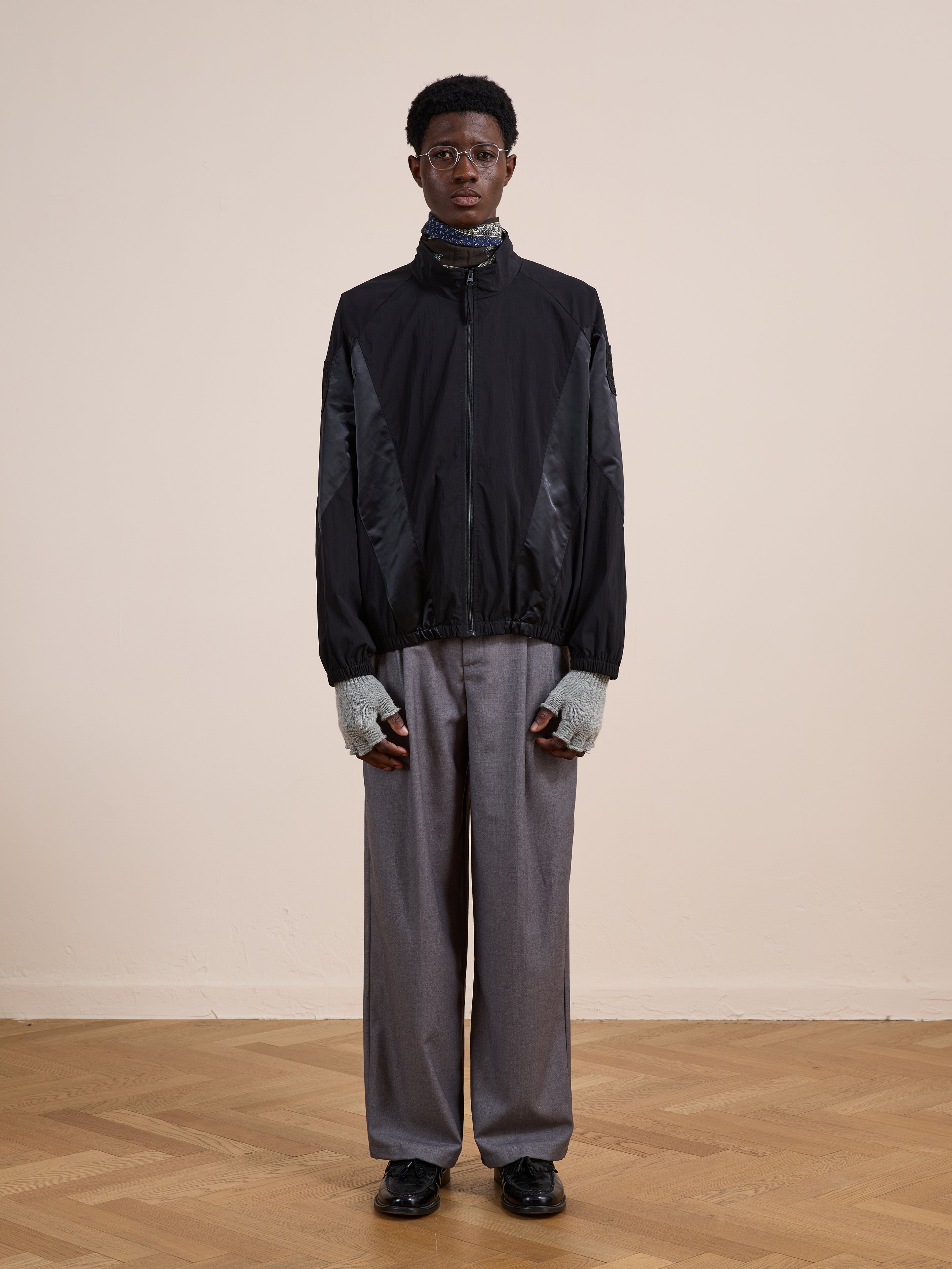 A person stands indoors against a plain wall, wearing the FOUND Pine and Rose Jacket, gray wide-leg trousers, gray fingerless gloves, black shoes, and glasses.