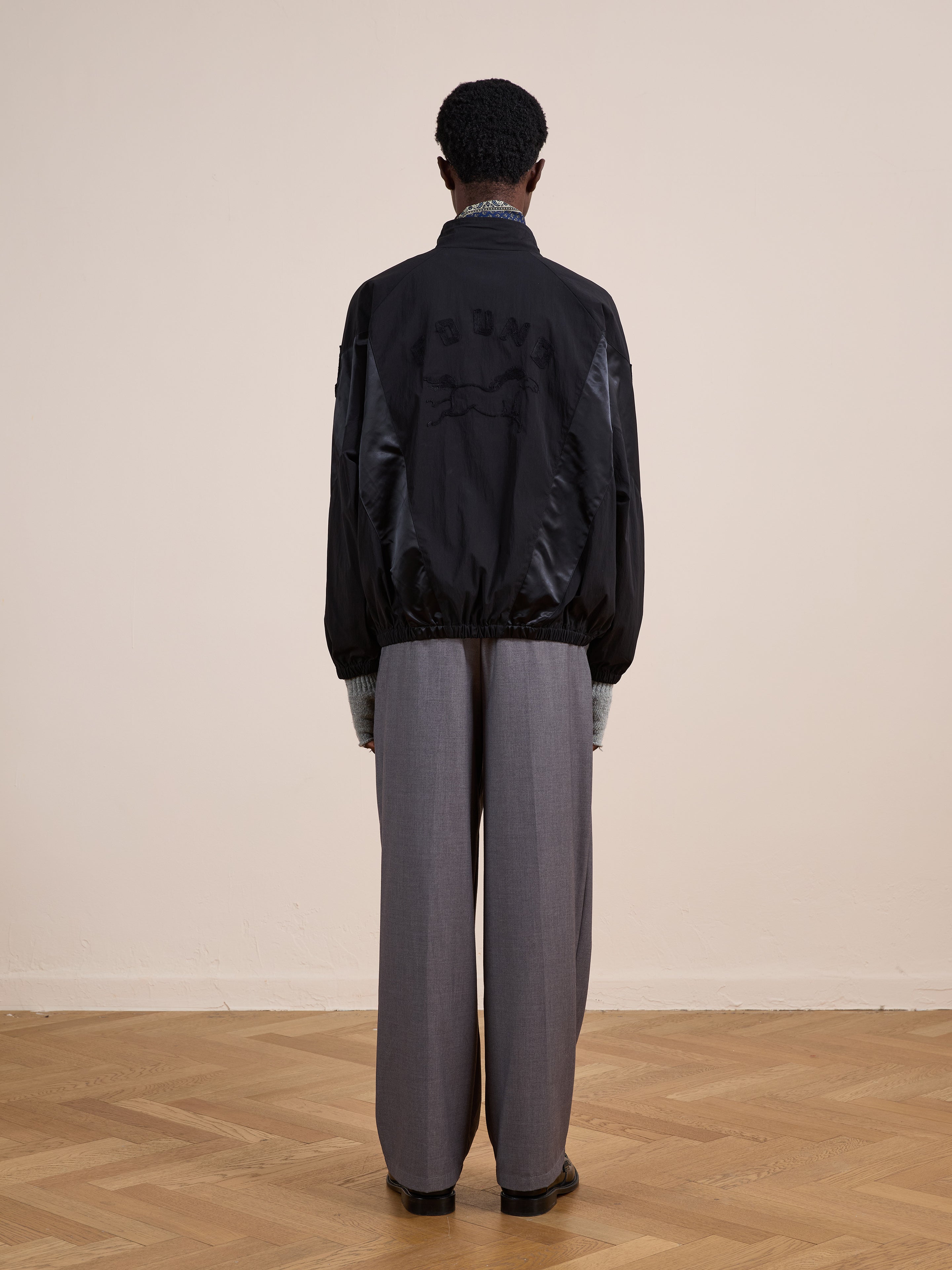 A person stands indoors on a wooden floor, facing away from the camera, wearing FOUND's Pine and Rose Jacket in black with wide-leg gray trousers—a stylish nod to vintage sportswear.