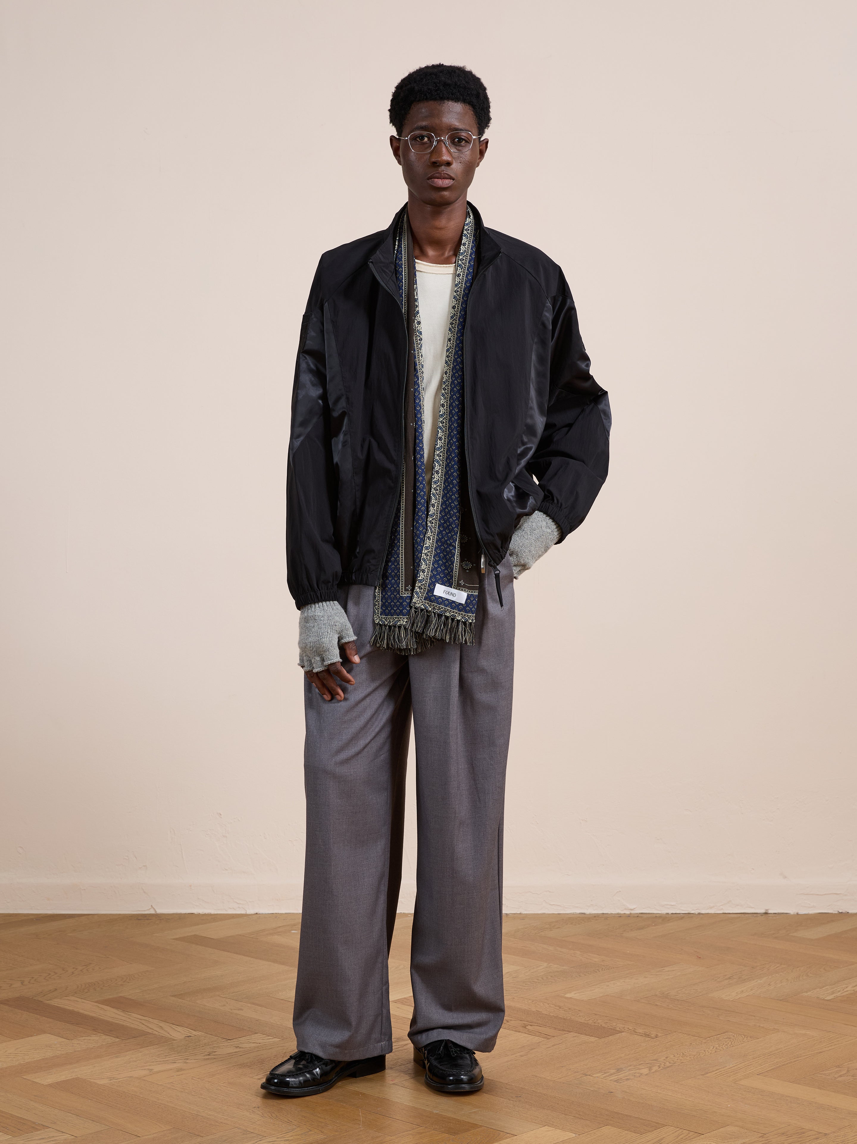 Indoors on a wooden floor, a person wears the FOUND Pine and Rose Jacket, light shirt, gray scarf, gray wide-leg trousers, black shoes, and glasses.