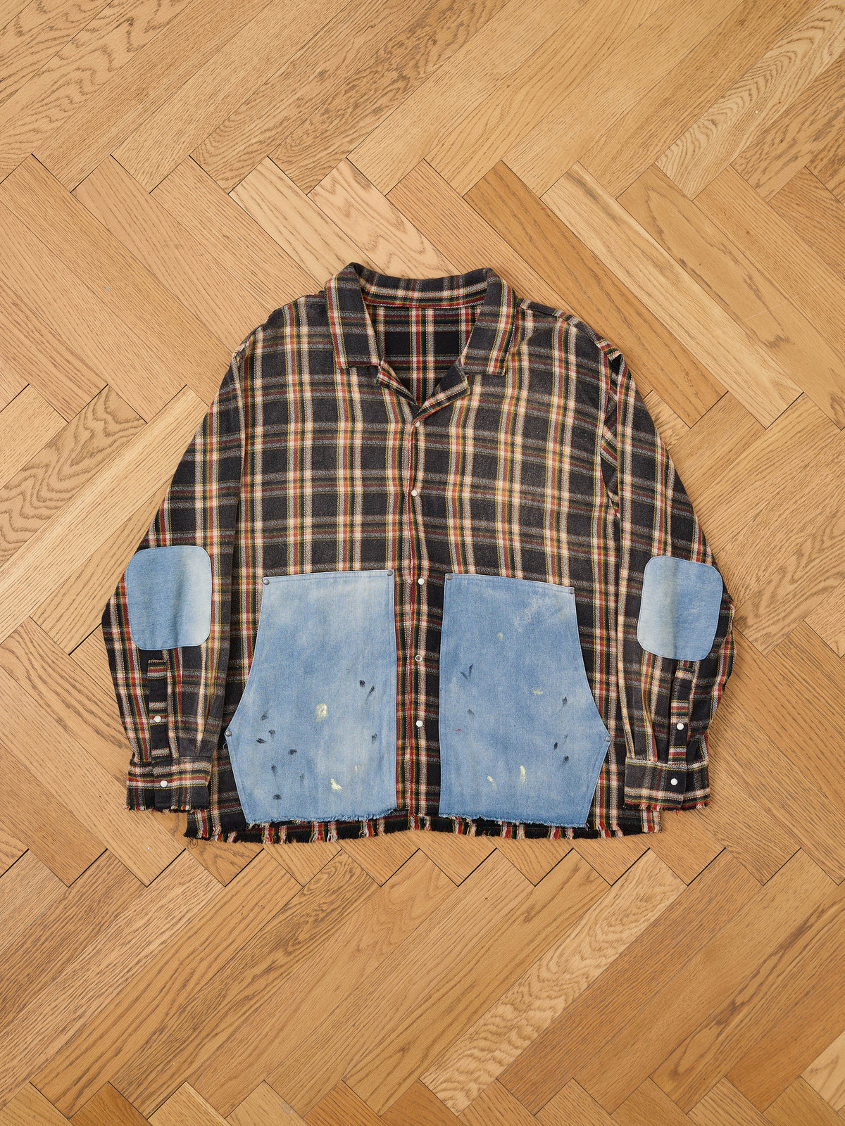 The FOUND Plaid Denim Shirt is a unisex relaxed fit with large denim patches on the elbows and lower front, laid flat on a light wooden herringbone floor.