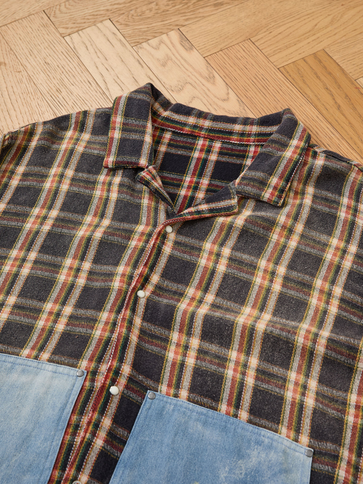 The FOUND Plaid Denim Shirt, a unisex relaxed fit with distressed denim pockets, is displayed flat on a wooden floor.