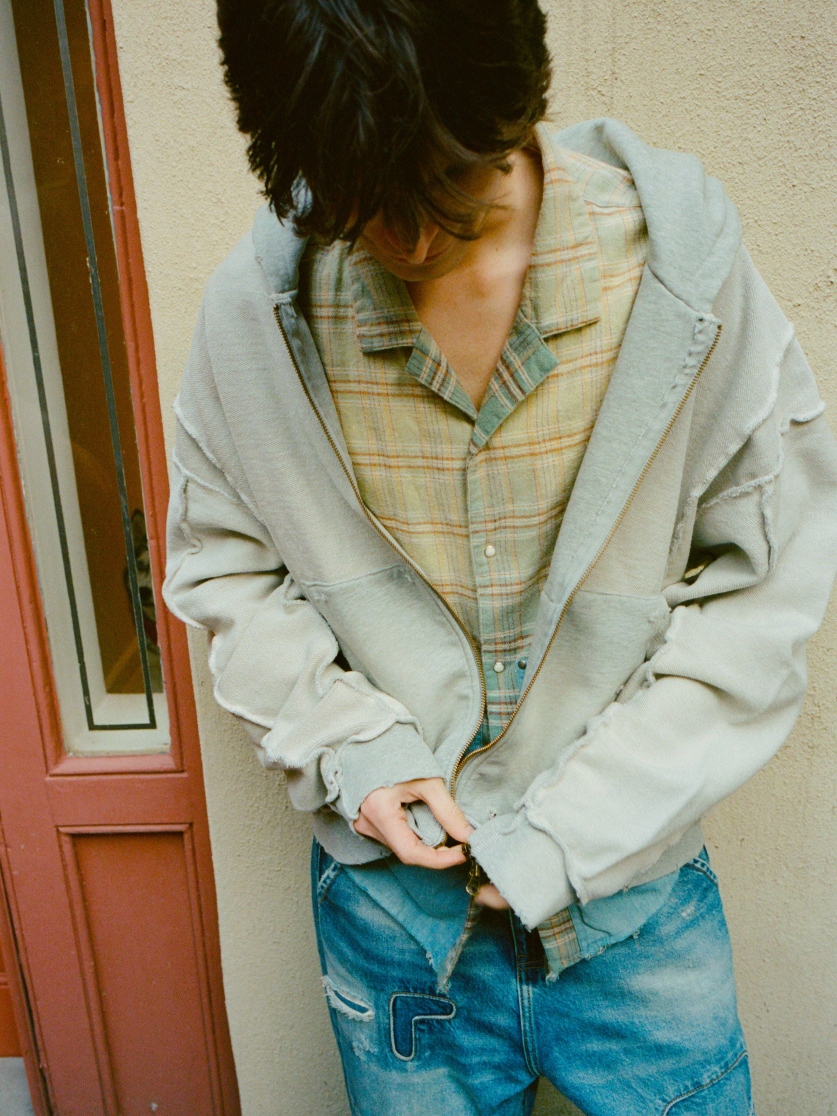 Wearing the FOUND Plaid Denim Shirt, a person in blue jeans adjusts their hoodie while looking down in front of a beige wall and red door.