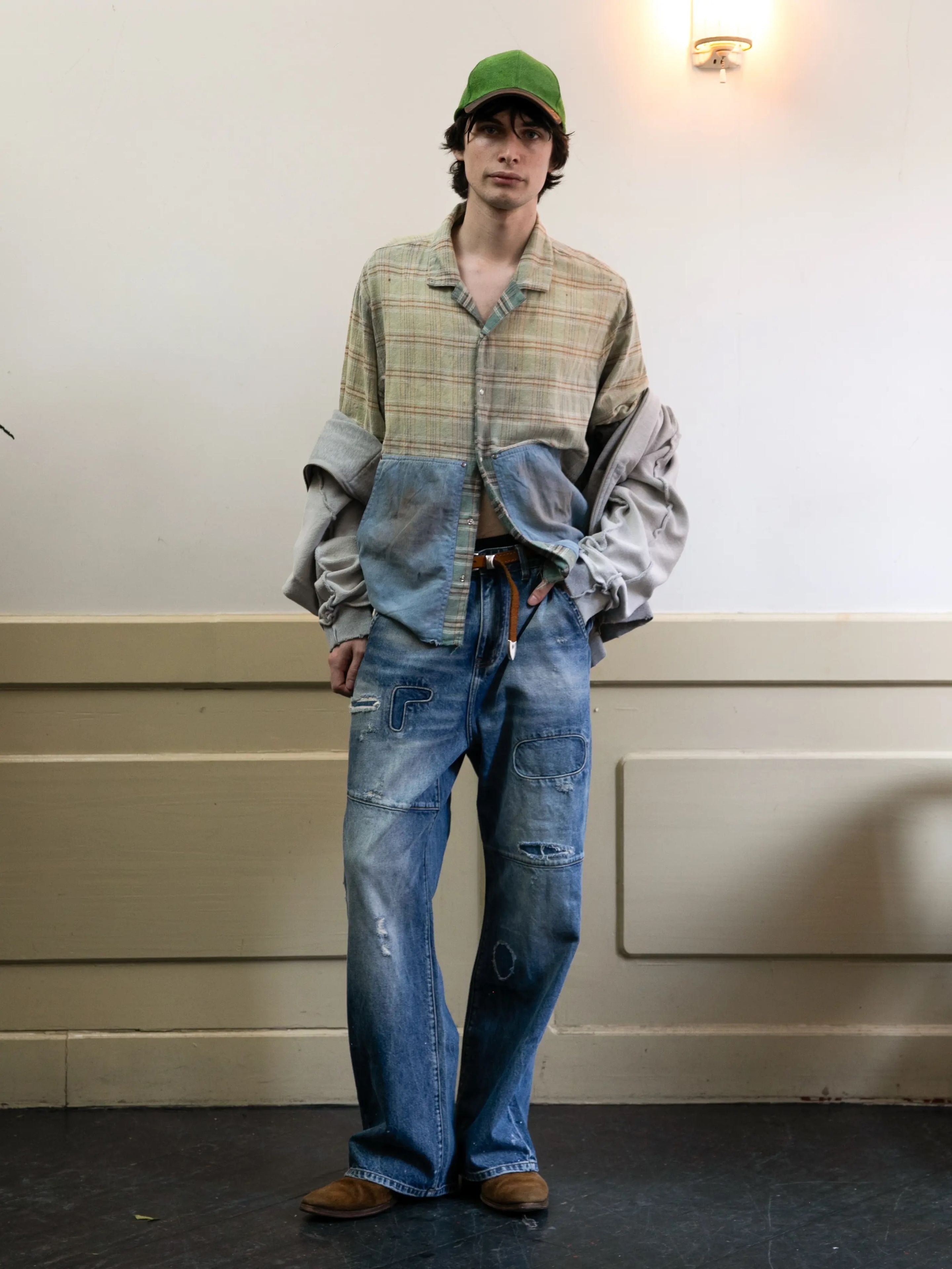 A person wearing the FOUND Plaid Denim Shirt with oversized sleeves, baggy distressed jeans, a green cap, and brown shoes stands indoors against a beige wall.