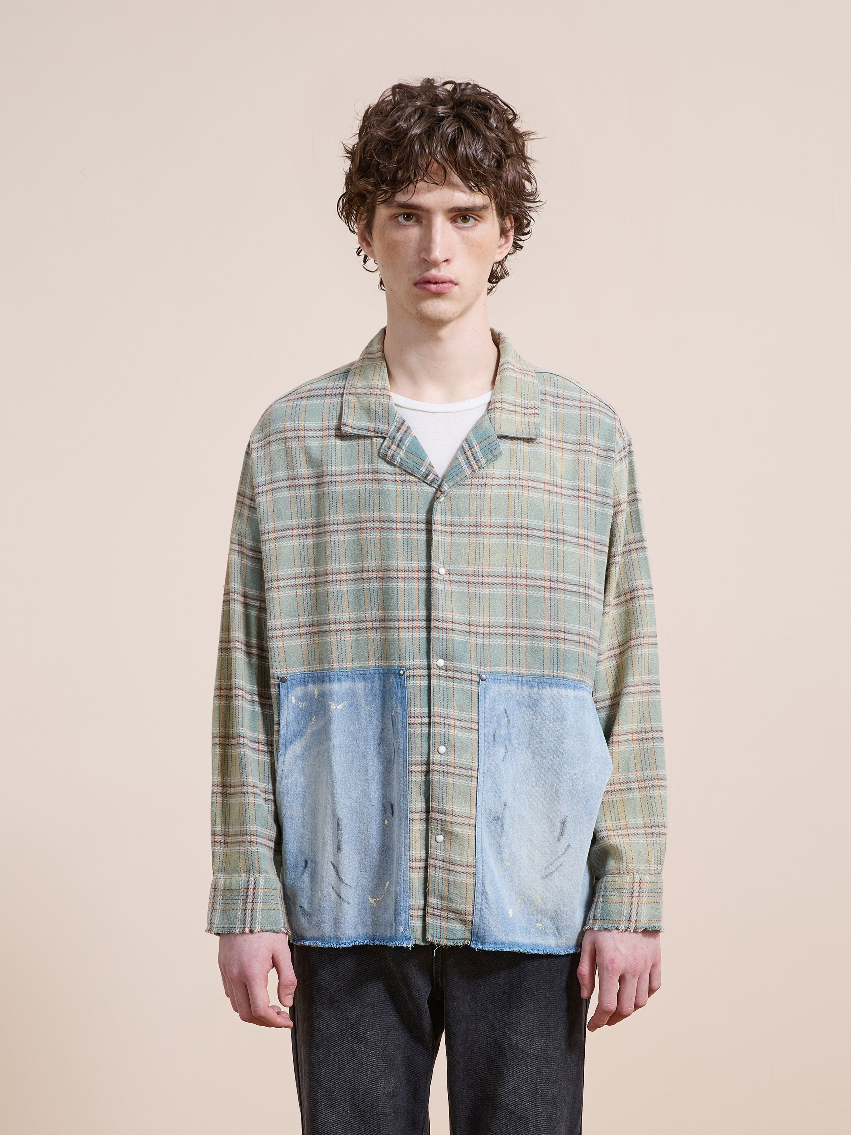 A young man stands facing forward in the FOUND Plaid Denim Shirt, a unisex relaxed-fit faded green plaid top with carpenter pockets, styled over a white t-shirt and dark pants against a plain beige background.