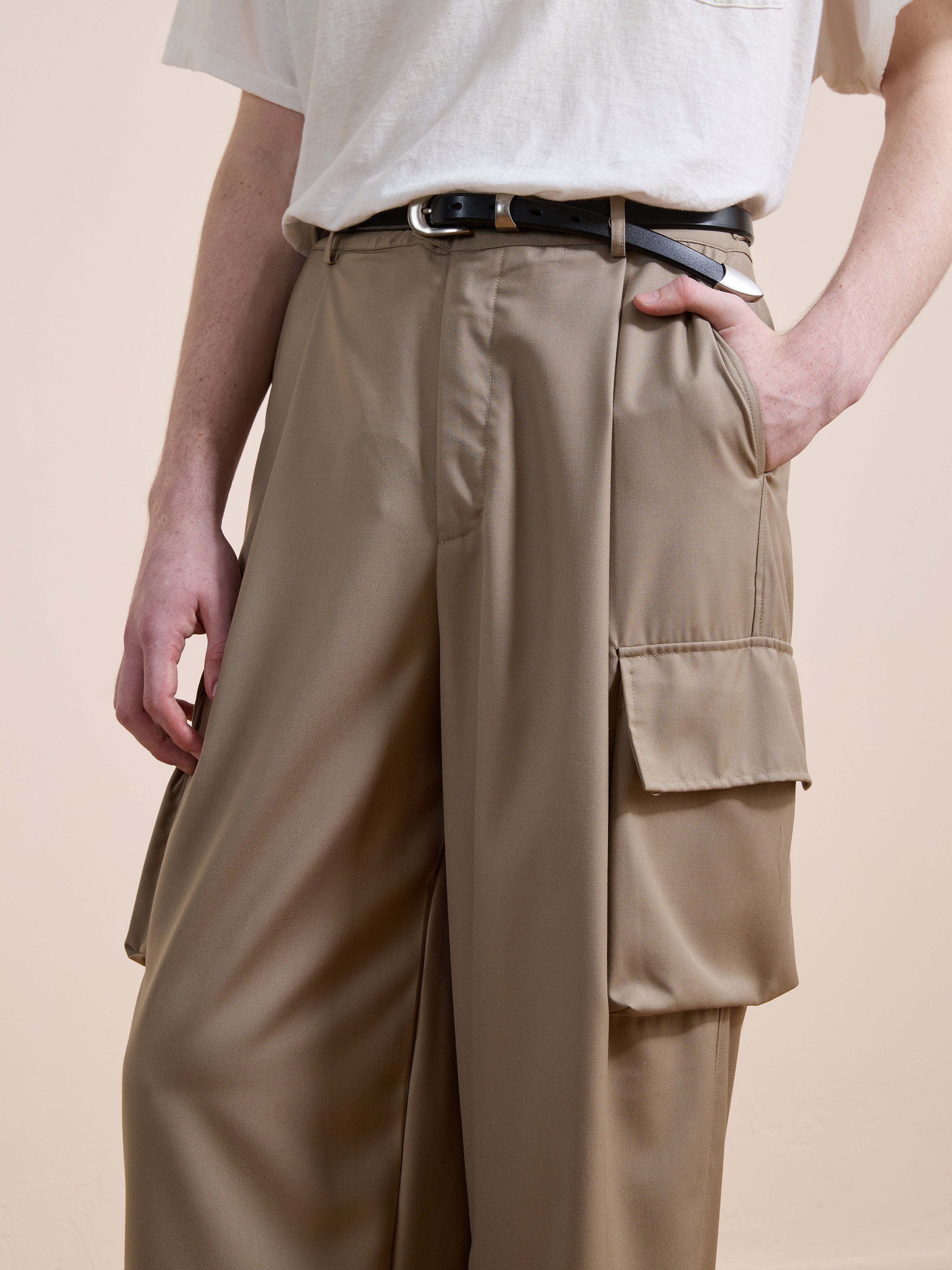 A person wears FOUND Pleated Cargo Pants with a relaxed fit, high waist, beige color, large pockets, and pleats, paired with a tucked-in white t-shirt and a black belt.