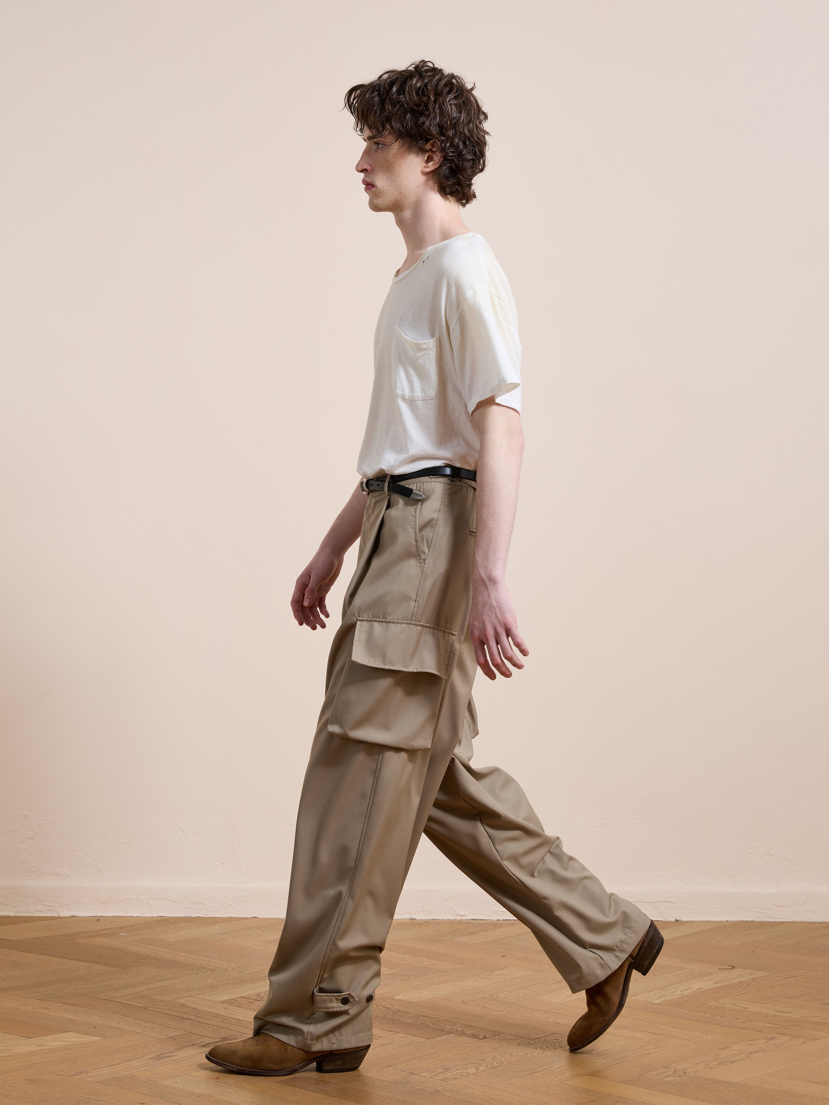 A person with curly hair walks indoors, wearing a white t-shirt, FOUND Pleated Cargo Pants in beige, a black belt, and brown shoes against a plain, neutral background.