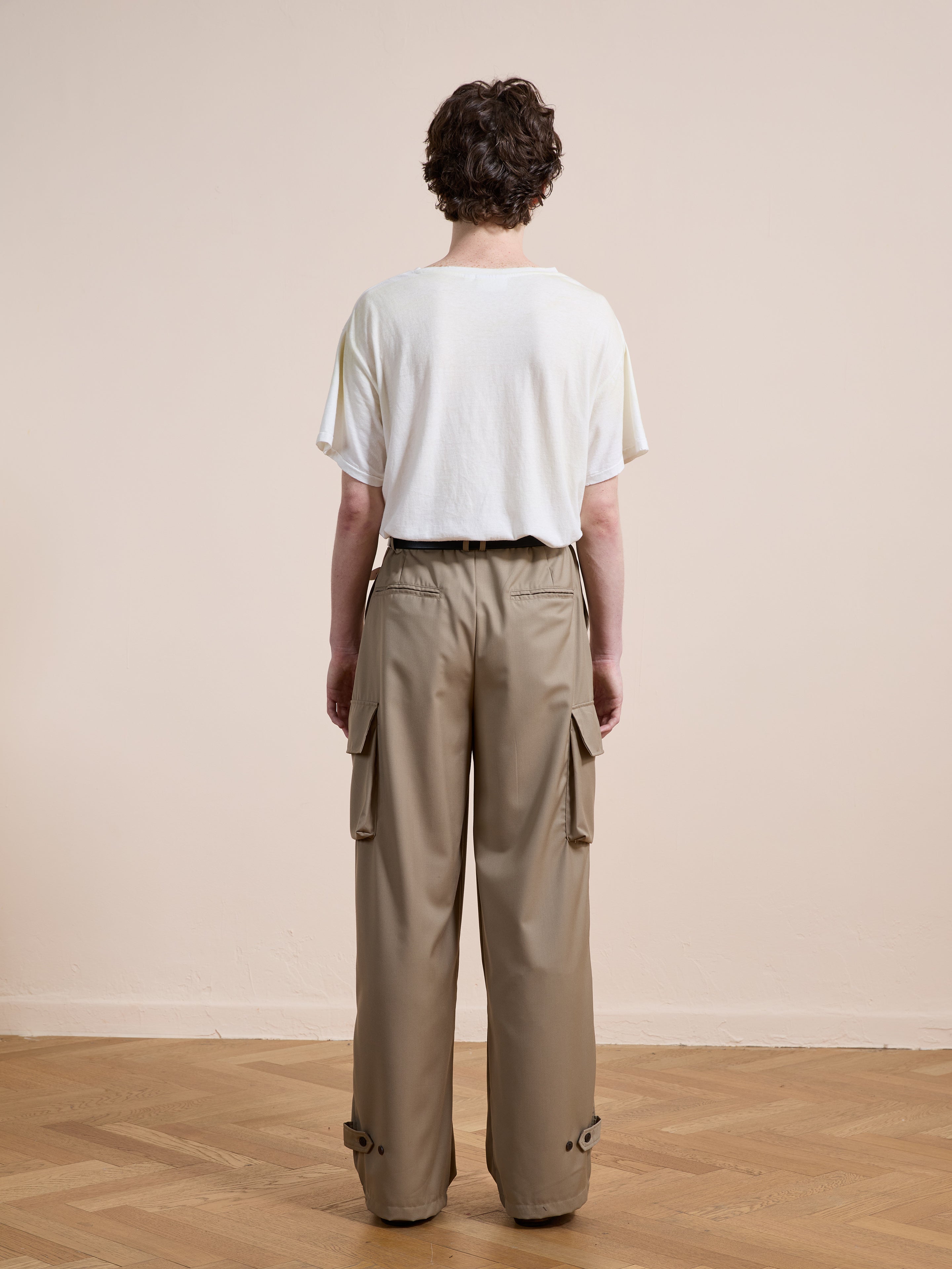 A person stands indoors on a wooden floor with a light-colored wall, wearing FOUND Pleated Cargo Pants and a loose white t-shirt, facing away from the camera.