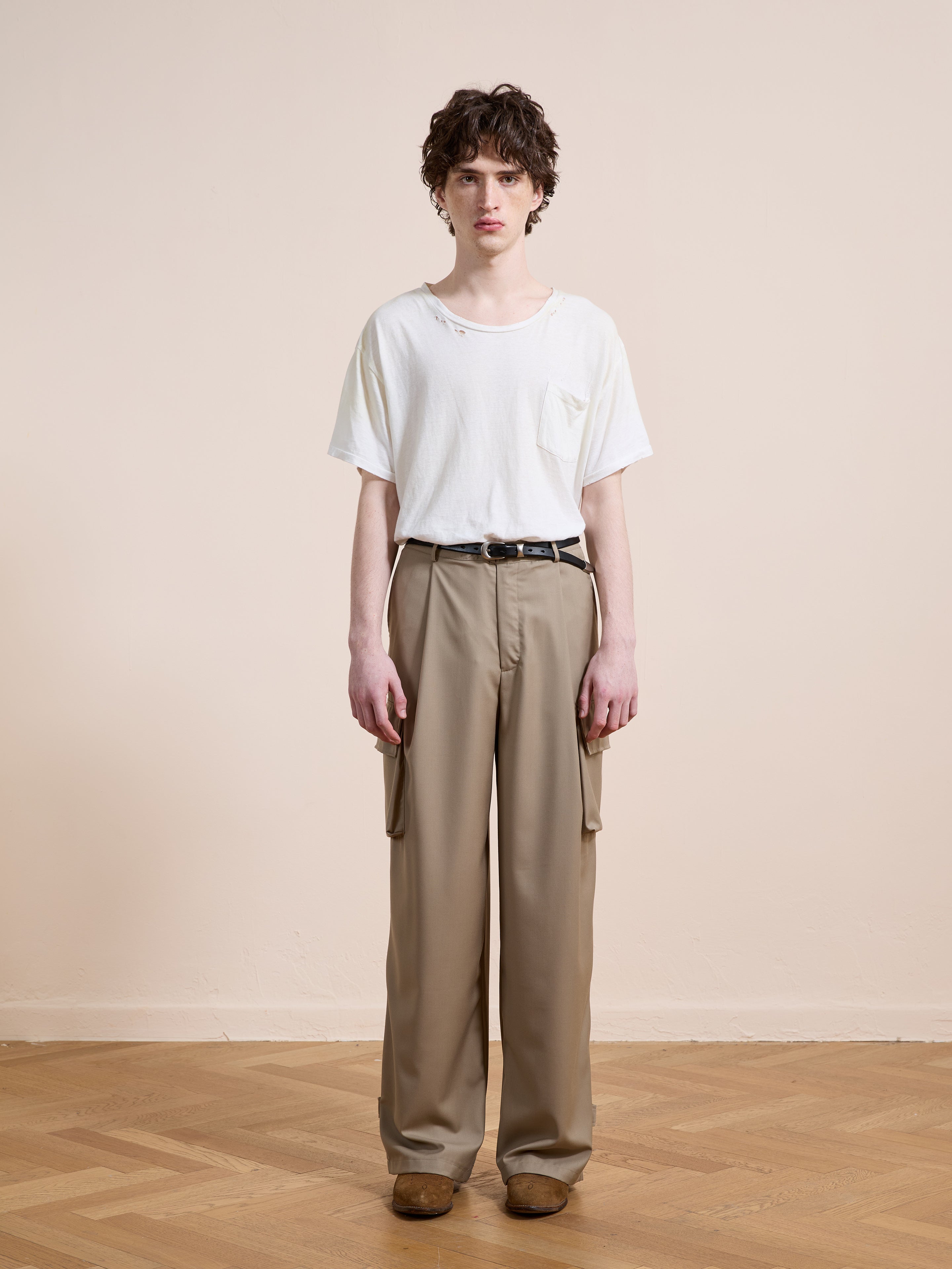A person stands indoors against a plain wall wearing a white t-shirt, FOUND Pleated Cargo Pants in beige with pockets, a black belt, and brown shoes.