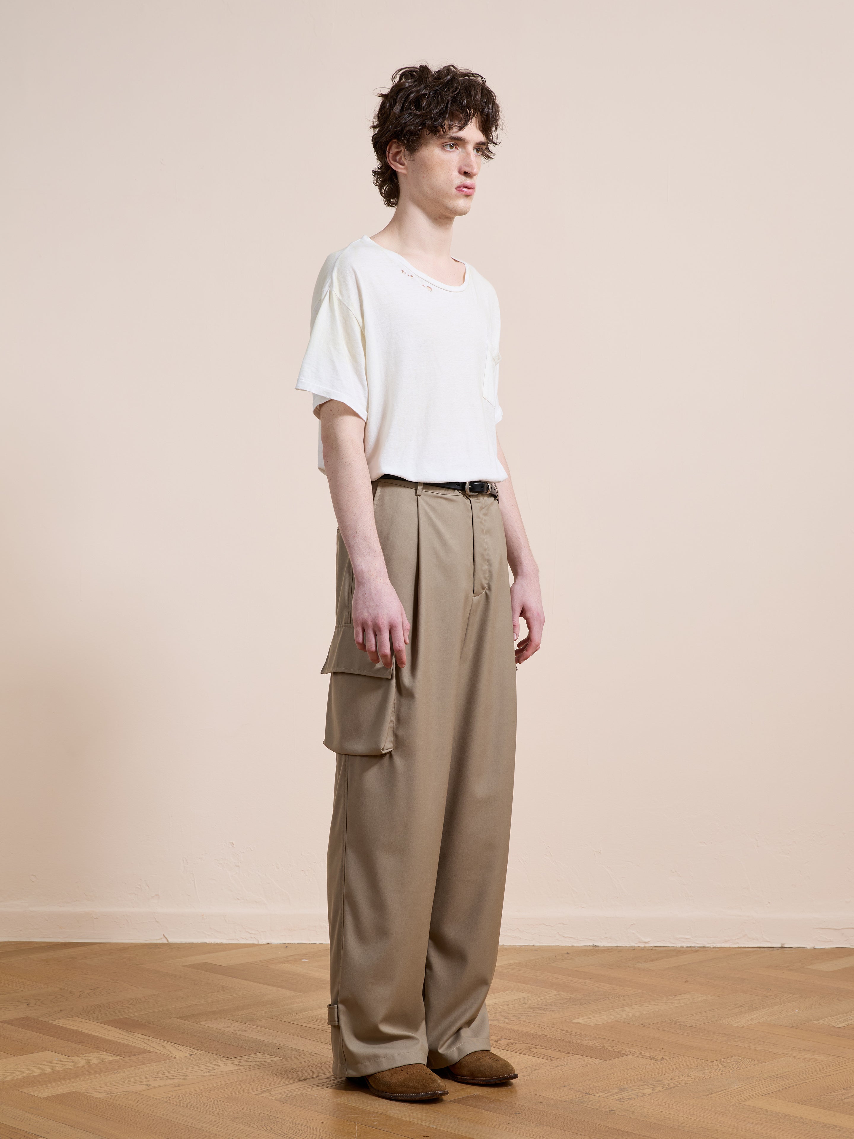 A person stands indoors against a light wall, wearing FOUND Pleated Cargo Pants in khaki with side pockets, paired with a loose white t-shirt and brown shoes.