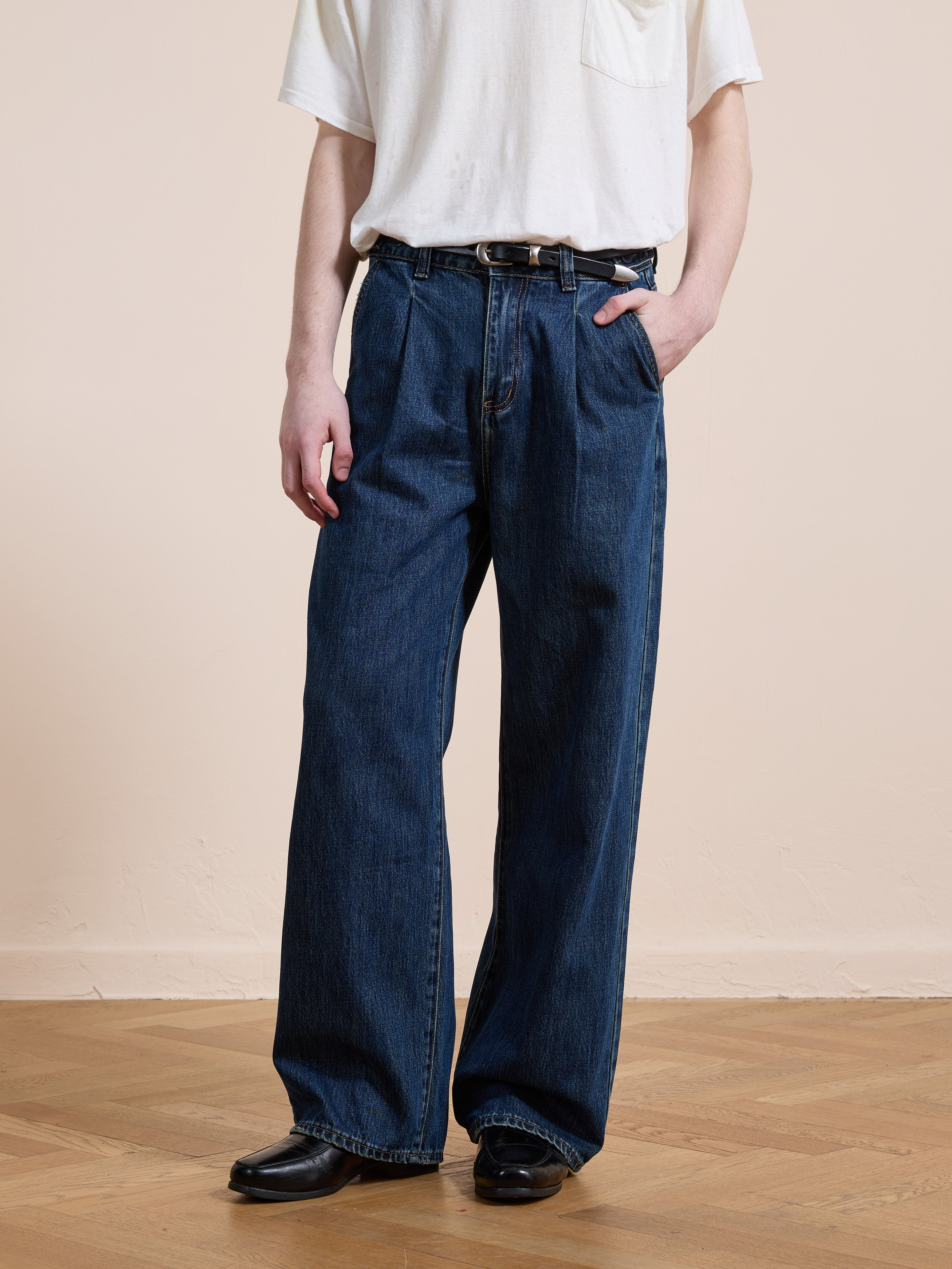A person wearing a white t-shirt, Found Pleated Relaxed Fit Jeans, and black shoes stands on a wooden floor with one hand in a pocket.