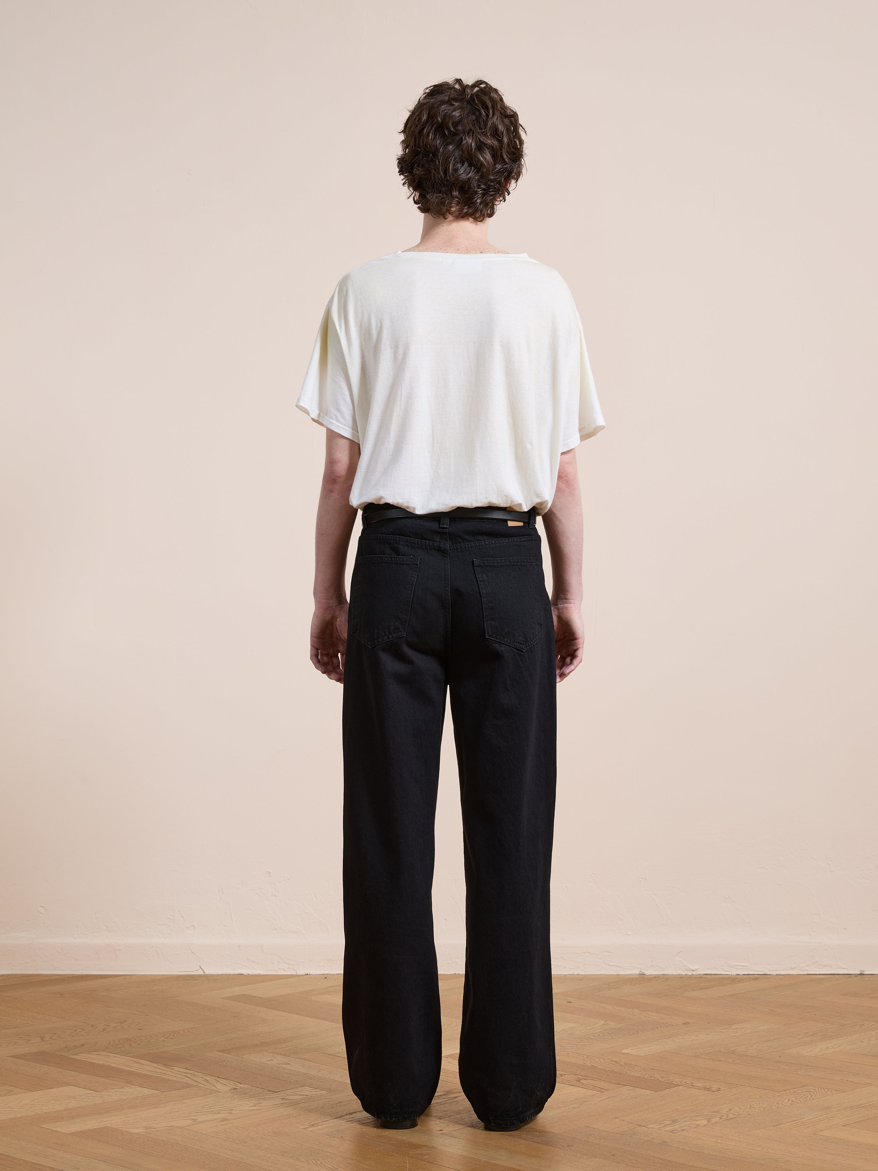 A person faces away on a wooden floor, wearing Found’s Pleated Relaxed Fit Jeans and a loose white T-shirt, set against a plain light-colored wall.
