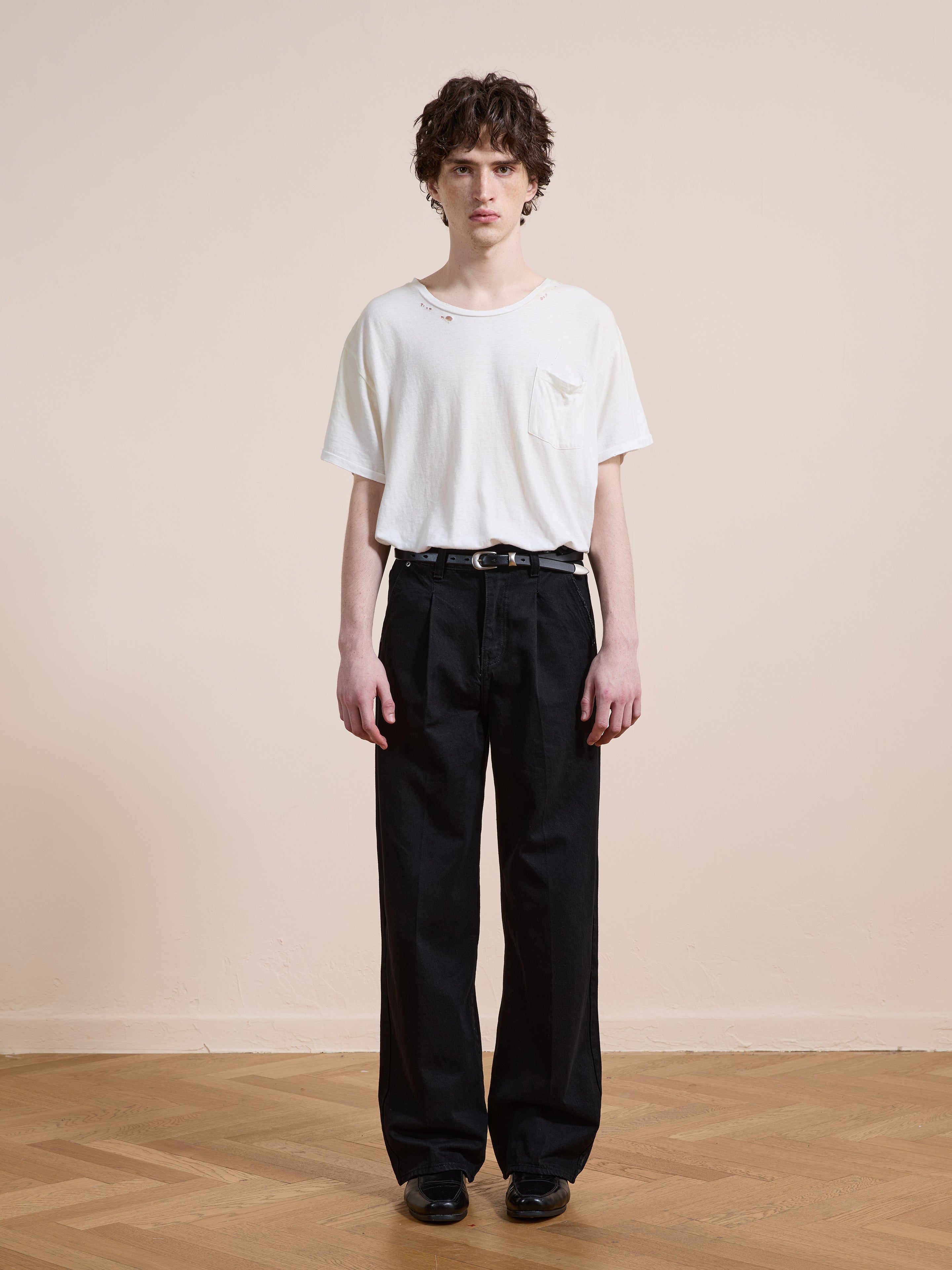 A person stands indoors against a plain wall, wearing black Pleated Relaxed Fit Jeans by Found and a white pocket t-shirt, looking straight at the camera.