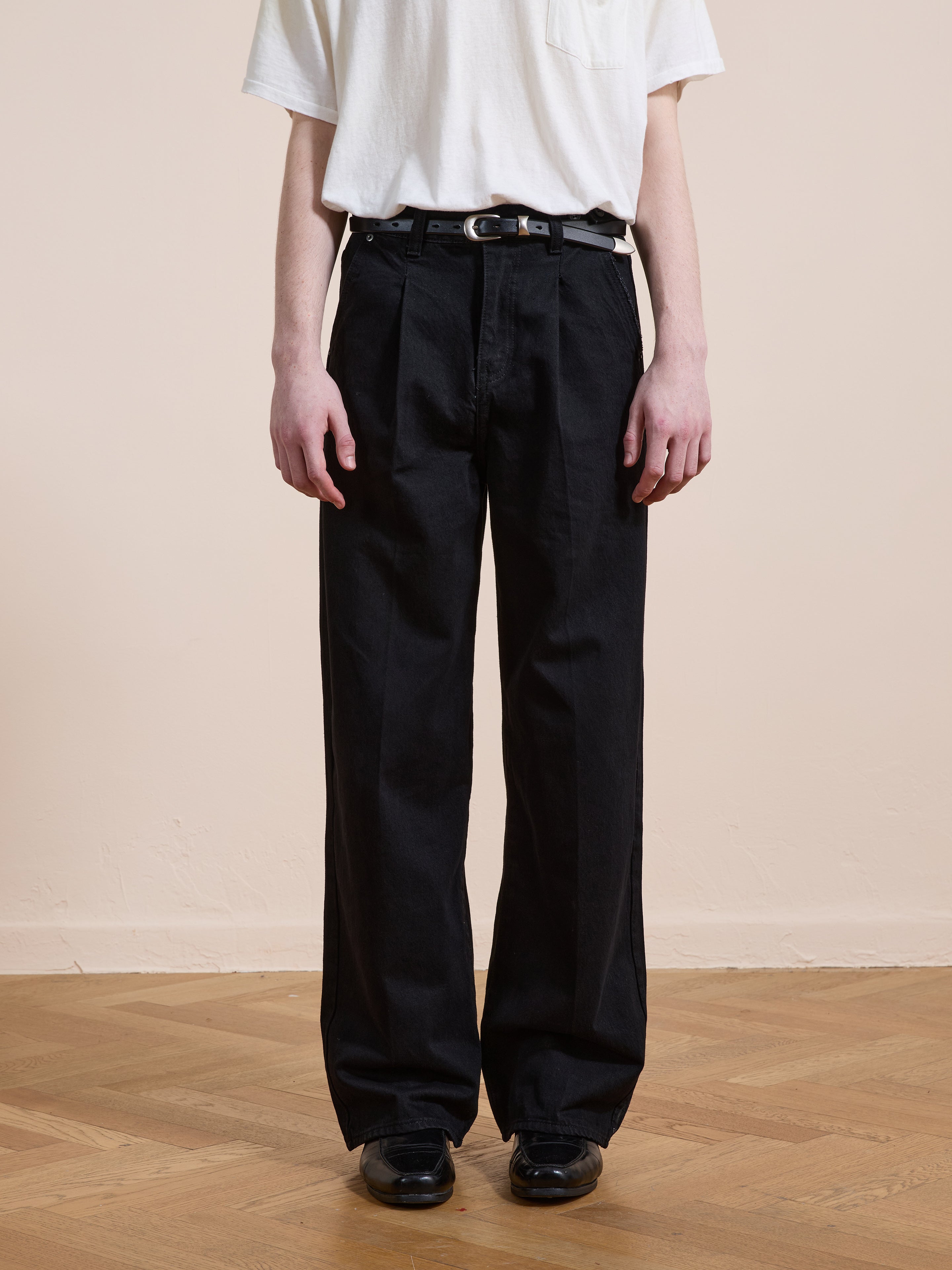 Model wears Found Pleated Relaxed Fit Jeans with a white t-shirt, black belt, and black shoes, standing on a wooden floor beside a beige wall.