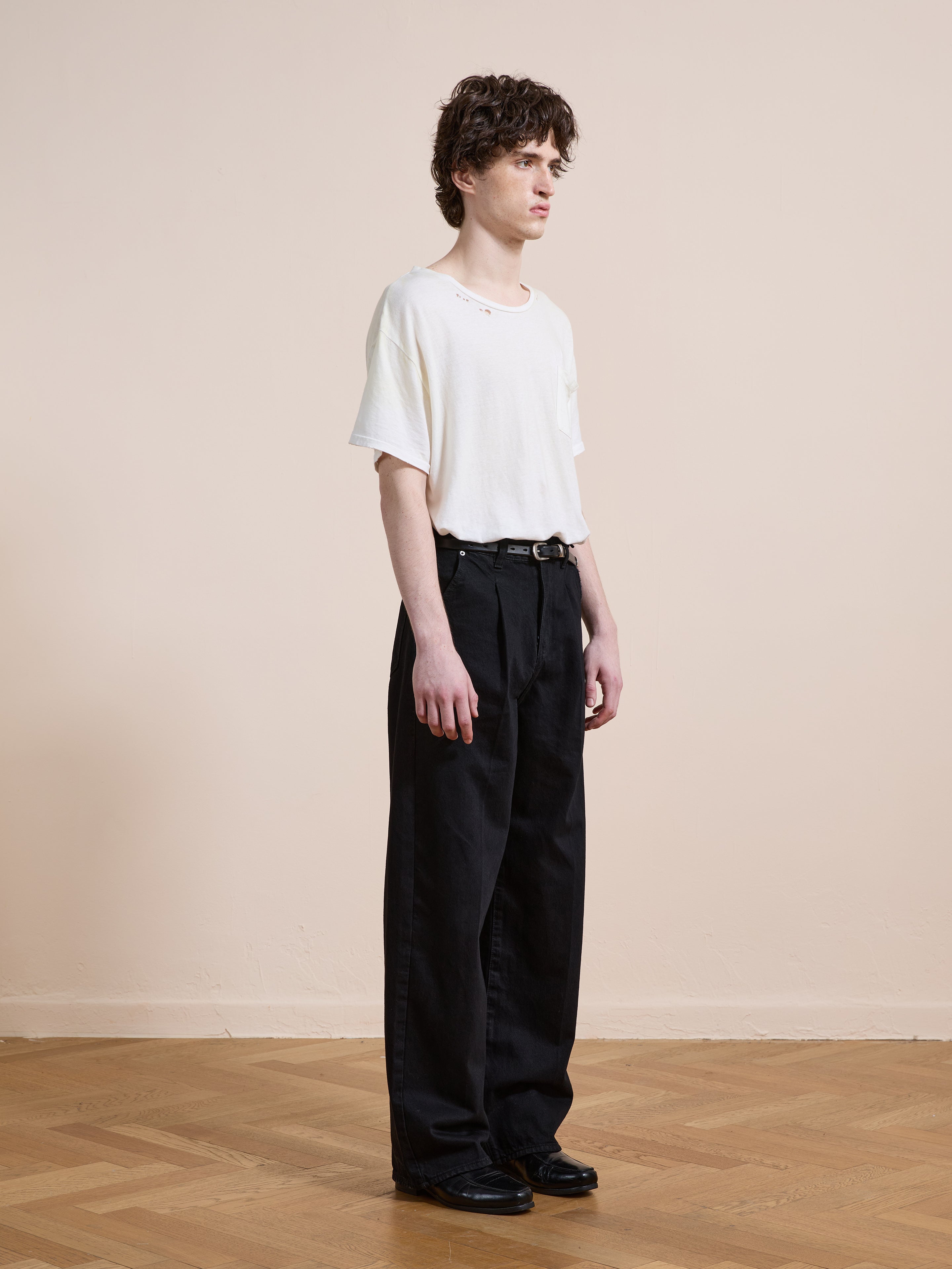 A person stands indoors on a wooden floor, facing sideways against a beige wall, wearing Found Pleated Relaxed Fit Jeans with a black belt, a white t-shirt, and black shoes.
