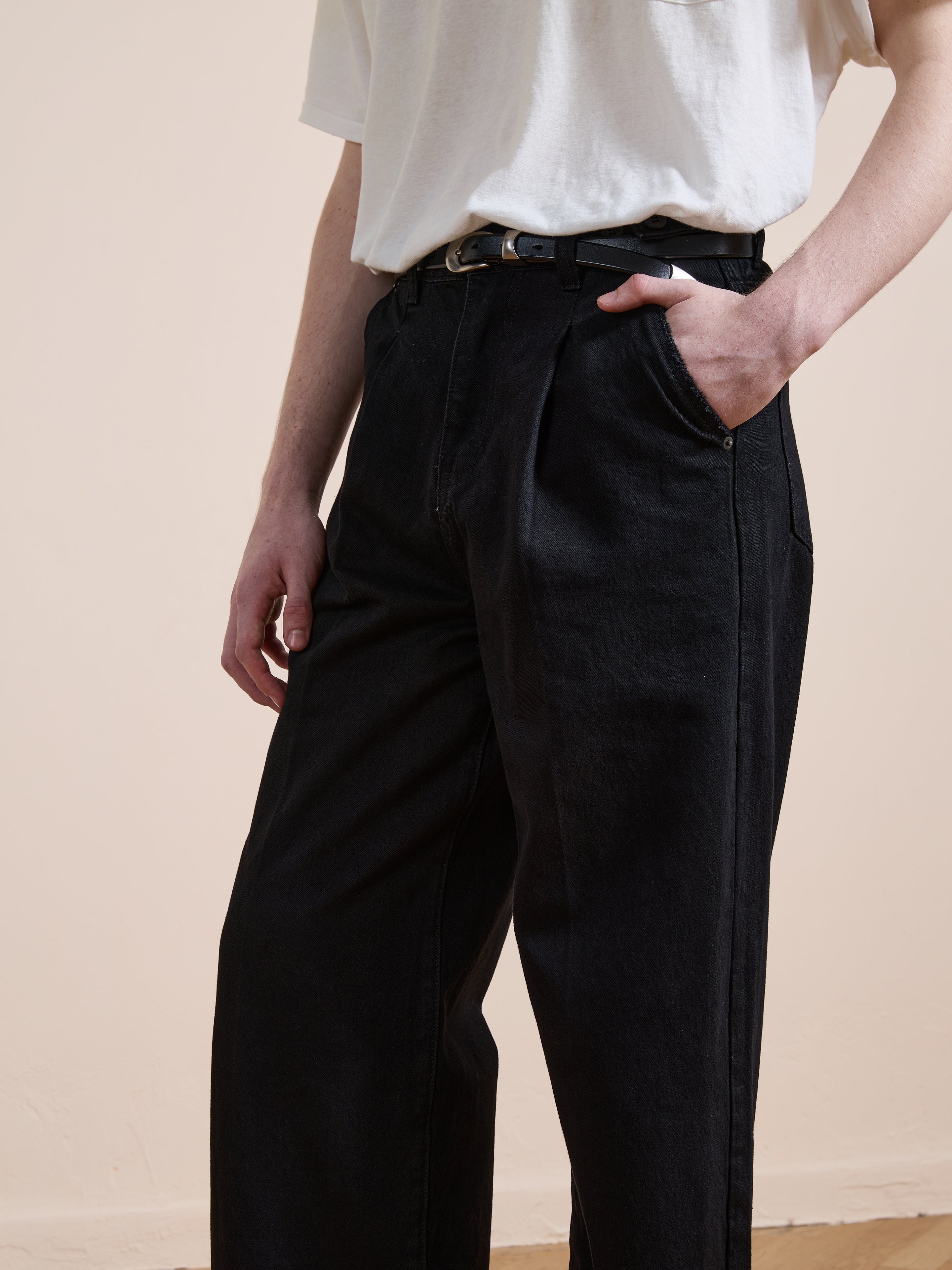 A person wears Found’s Pleated Relaxed Fit Jeans with a belt and a white t-shirt, one hand in the pocket, standing against a plain light background.