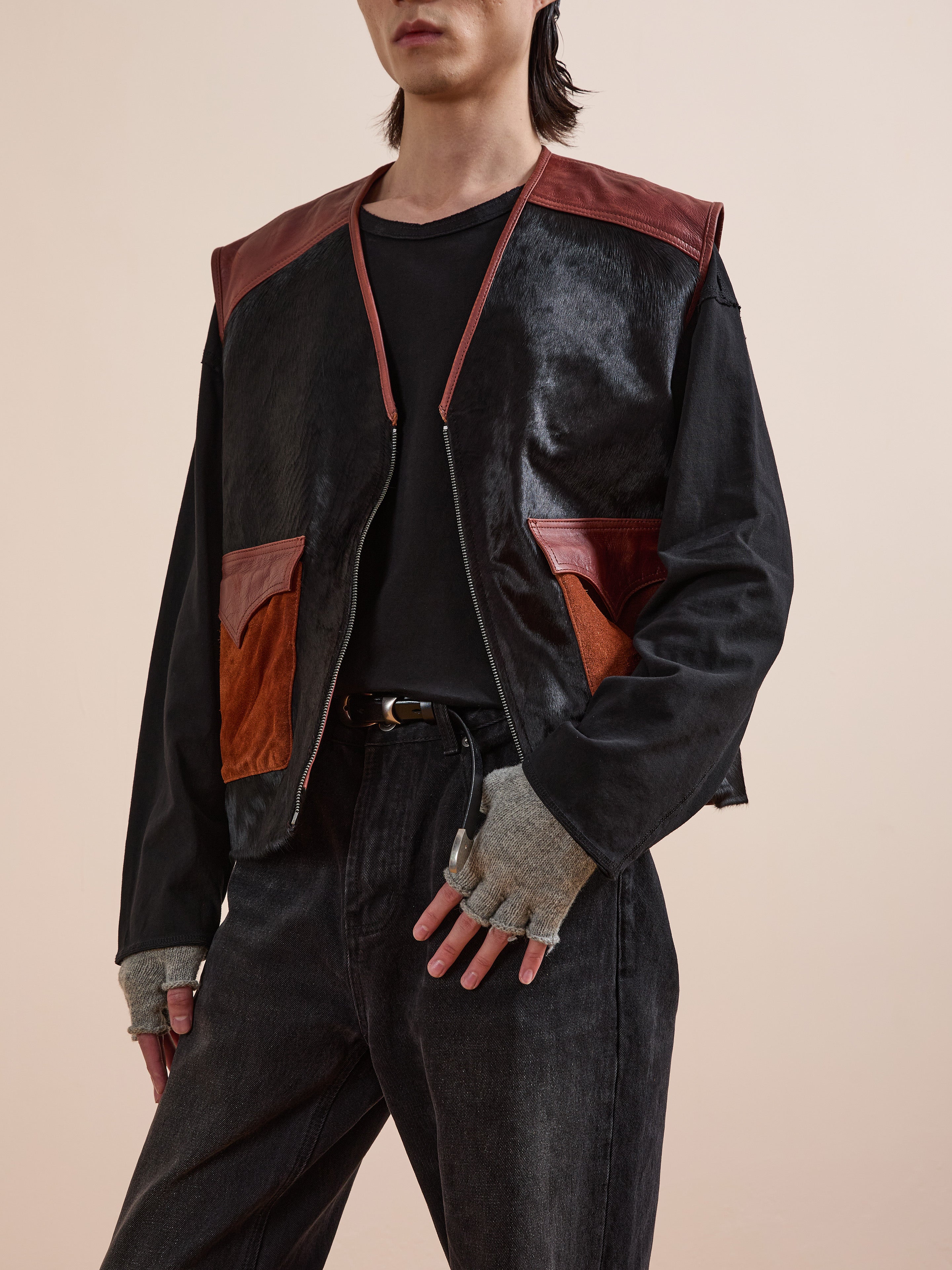 A person poses against a plain beige background wearing a black shirt, black jeans, Found’s Fur Western Leather Vest in black and brown, fingerless gray gloves, and a black belt.