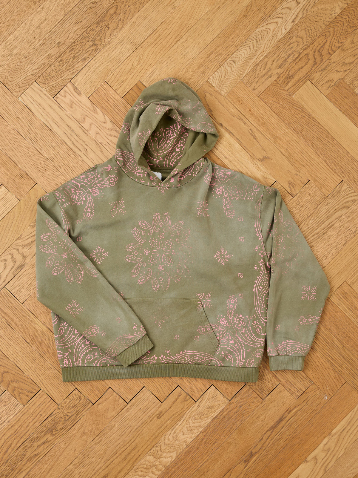 The FOUND Printed Paisley Hoodie, a green unisex pullover with a pink paisley pattern, is displayed flat on a wooden herringbone floor.