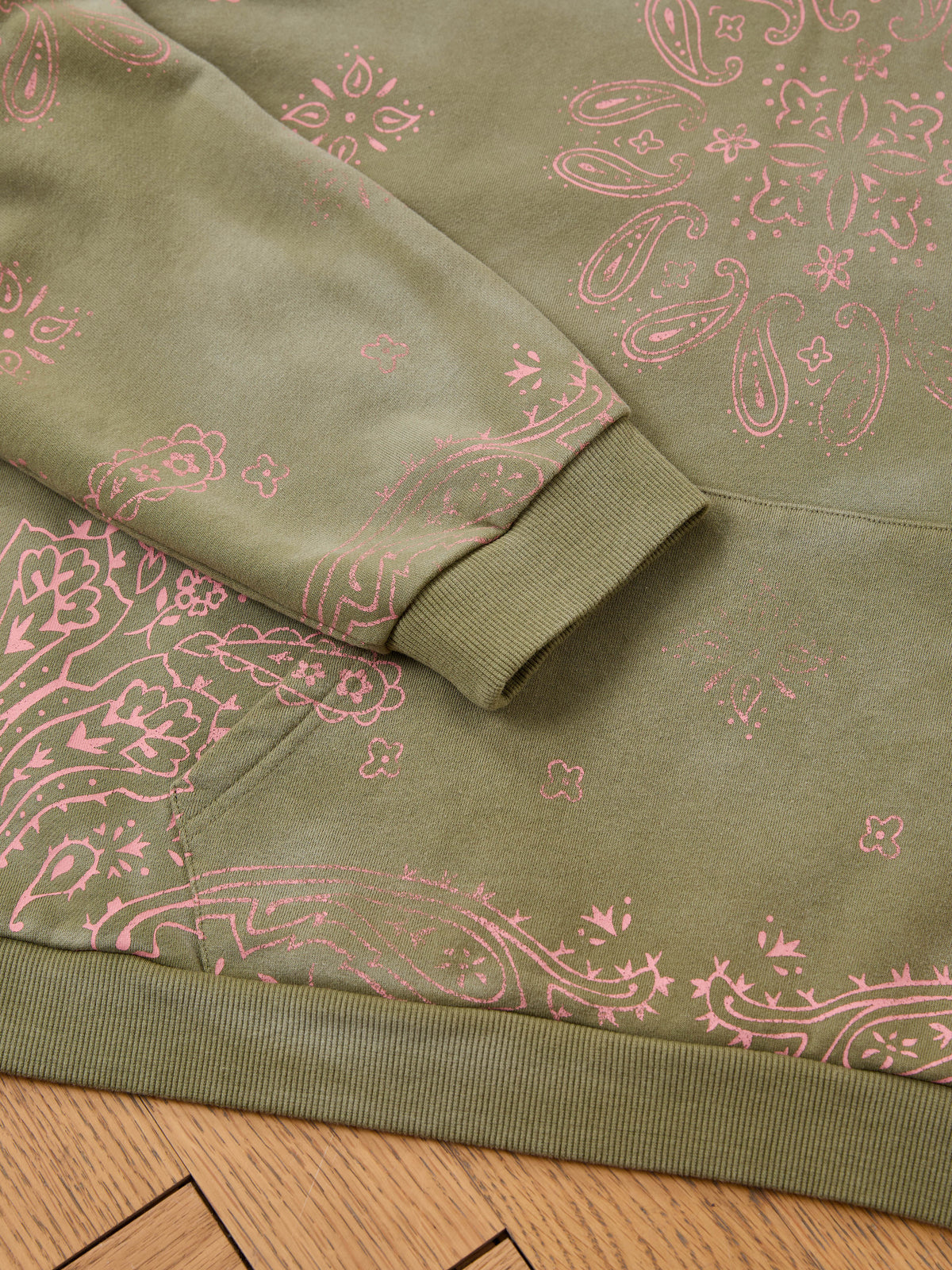 Close-up of FOUND Printed Paisley Hoodie with pink paisley on olive green, laid flat on wood—a stylish unisex pullover for any casual look.