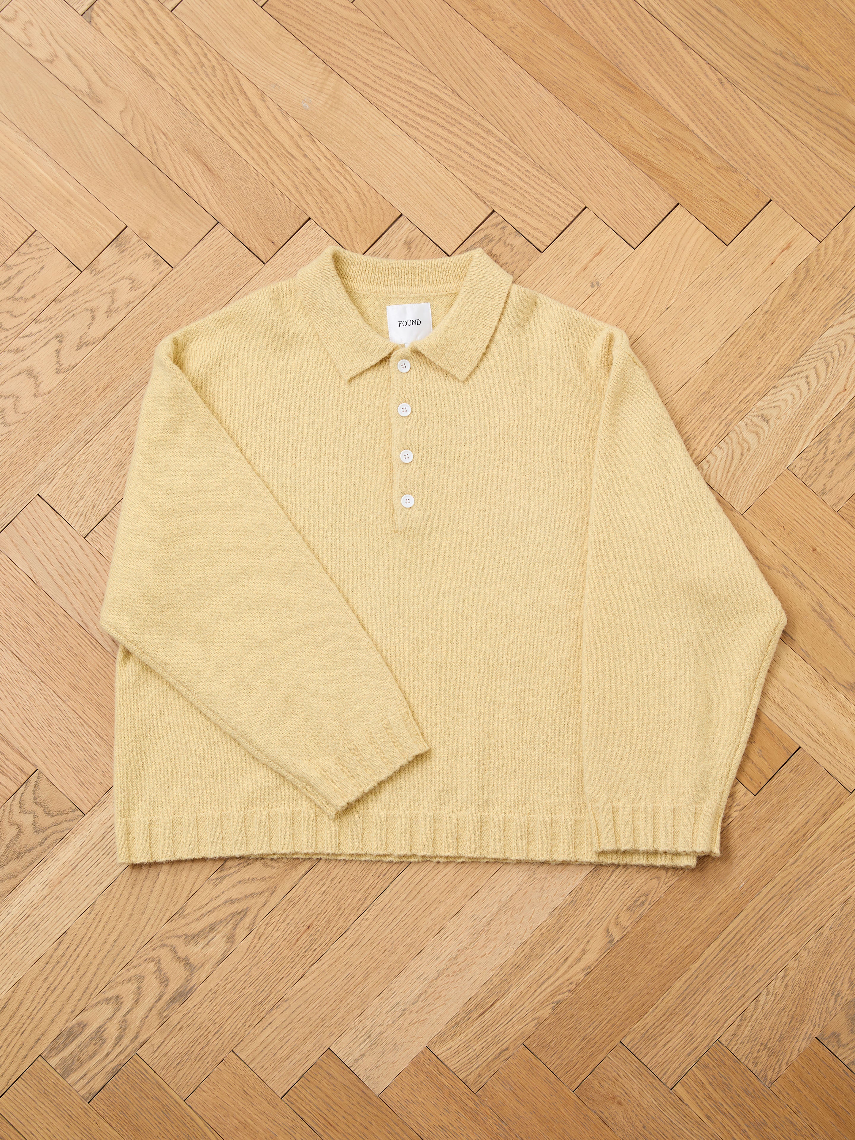 The FOUND Knit LS Polo Sweater in light yellow, made from soft brushed knit with buttons, is laid flat on a wooden herringbone-patterned floor.