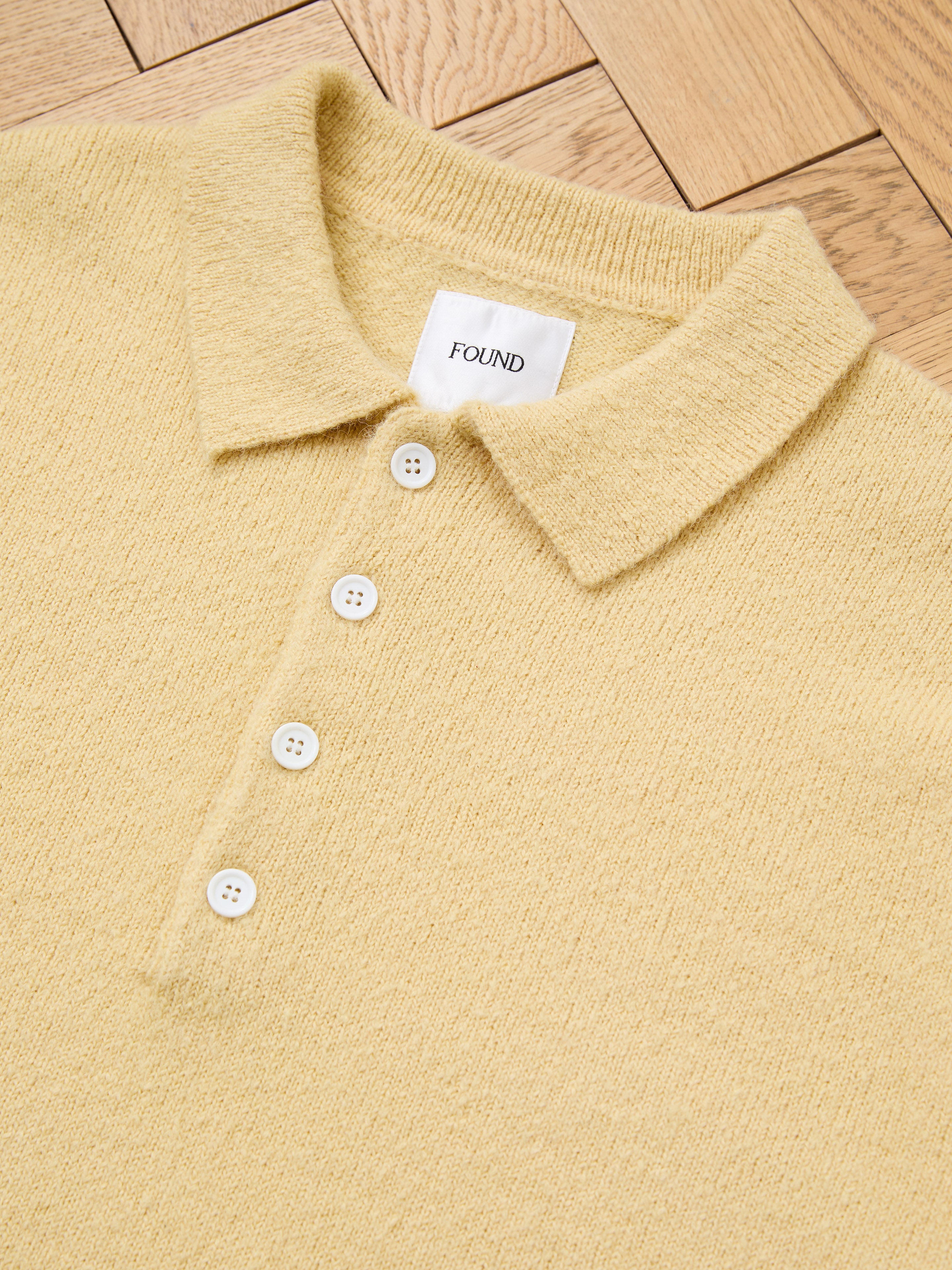 A yellow FOUND Knit LS Polo Sweater, featuring a textured collar, four white buttons, and a "FOUND" tag, made from soft brushed knit fabric and displayed flat on a wooden surface.