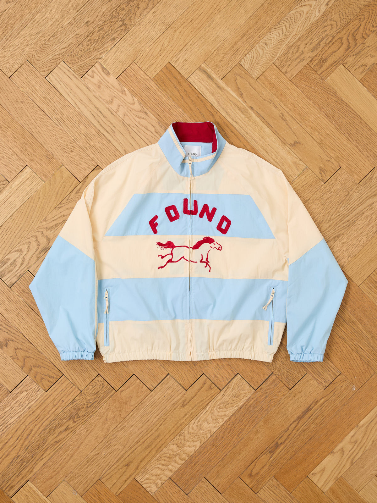 The Racing Striped Windbreaker by FOUND is a beige and light blue unisex lightweight jacket featuring red “FOUND” text and a running dog graphic on the front, reminiscent of vintage sportswear, displayed on a wood parquet floor.