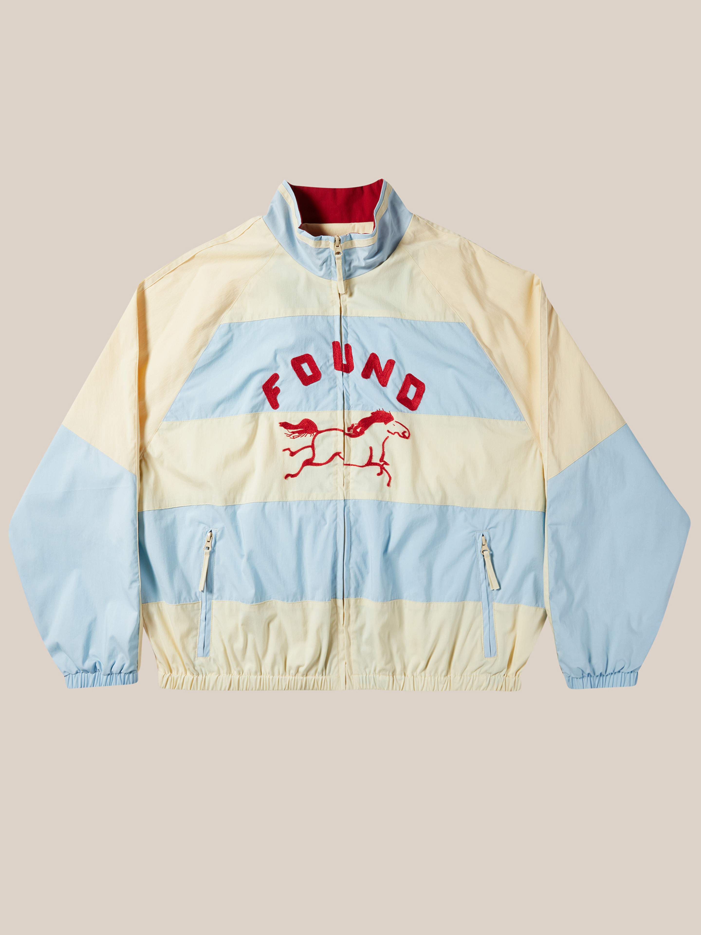 The FOUND Racing Striped Windbreaker is a unisex cream and light blue jacket inspired by vintage sportswear, with "FOUND" and a red racing horse embroidered on the front, zip closure, and two front pockets.
