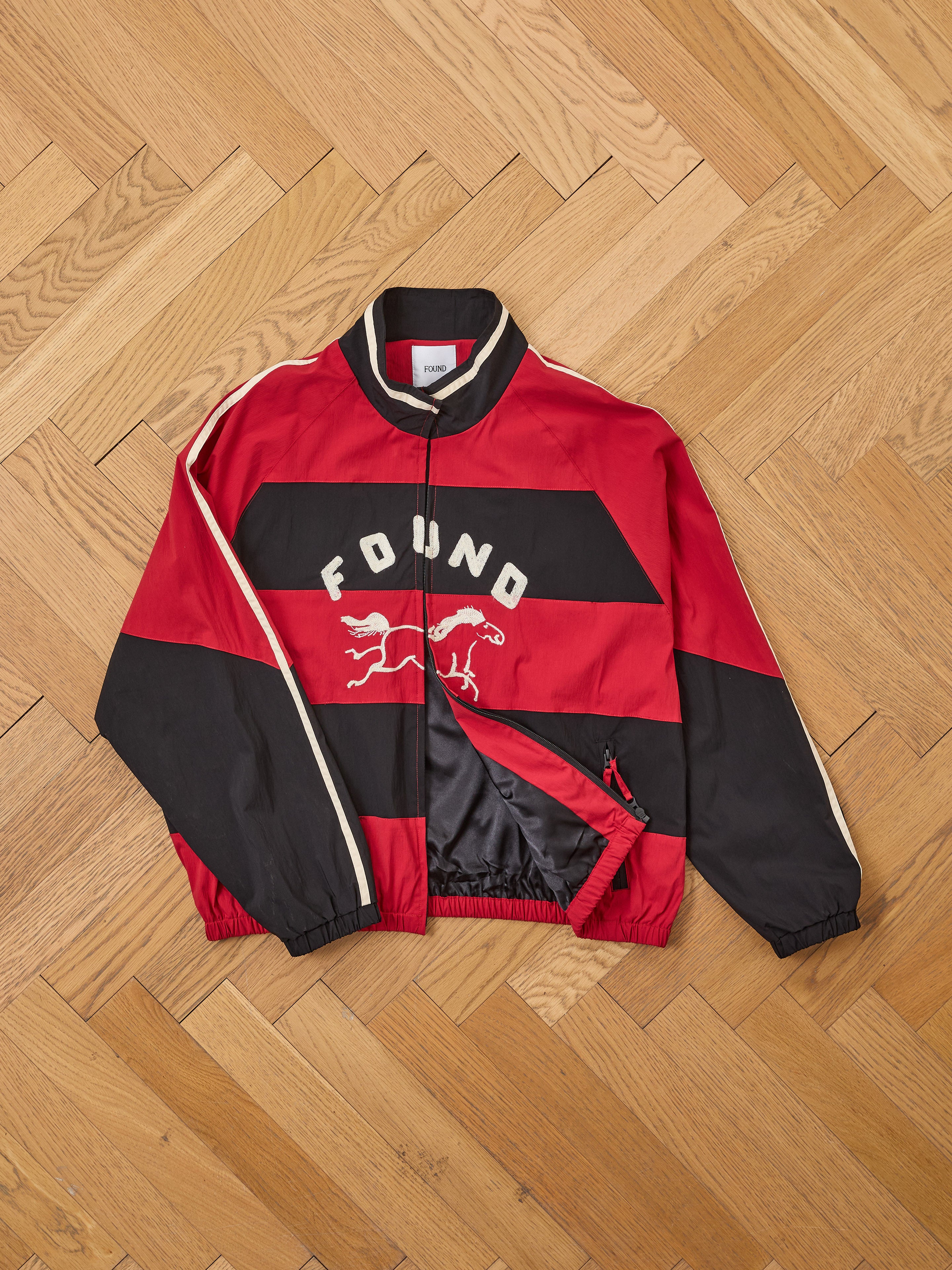 A FOUND Racing Striped Windbreaker in red and black with "FOUND" and a horse graphic on the front, displayed flat on a wooden herringbone floor—a striking vintage sportswear piece.