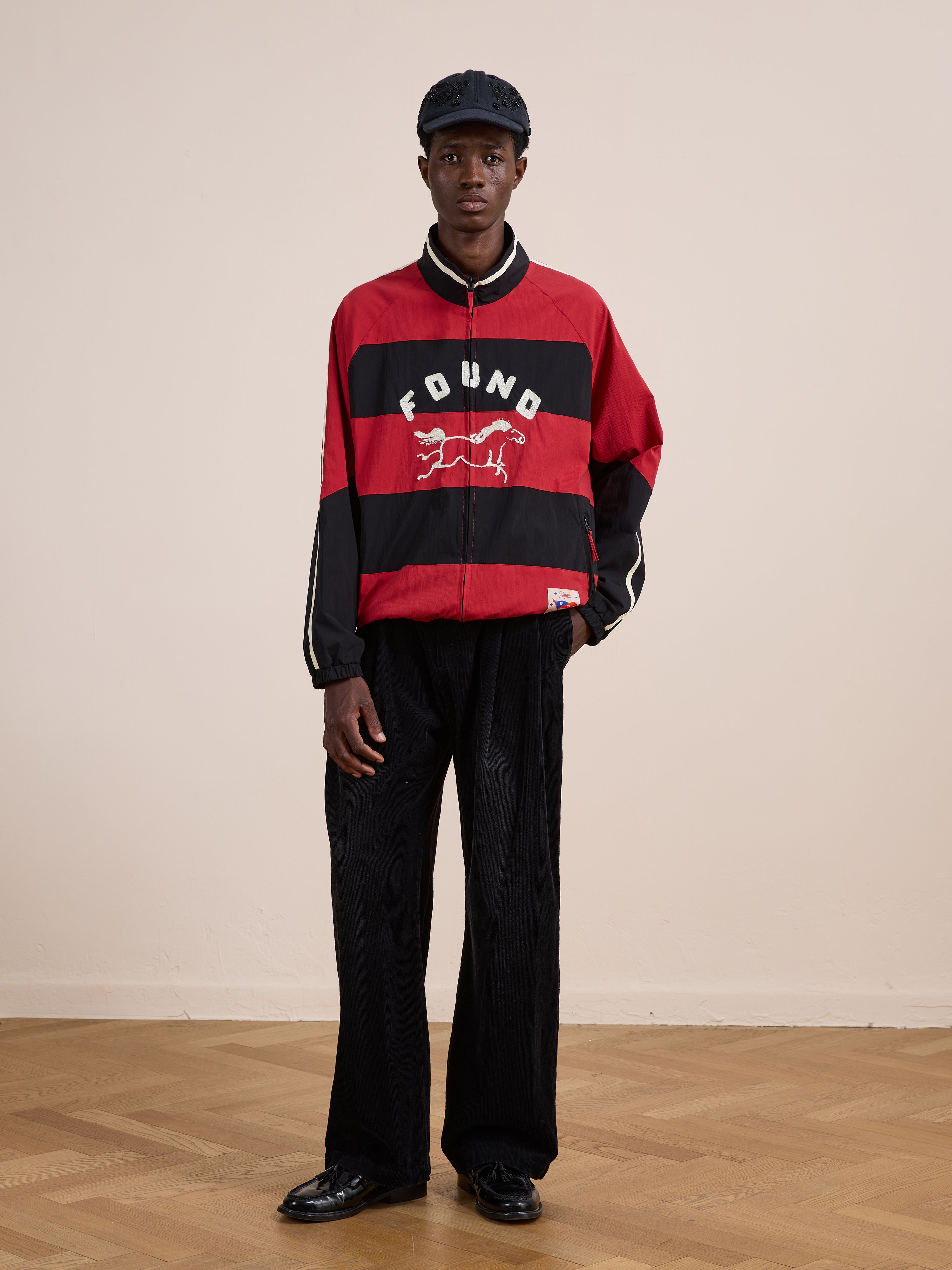 A person wears a black cap, FOUND Racing Striped Windbreaker, black wide-leg pants, and black shoes, standing indoors on a wood floor against a plain wall.
