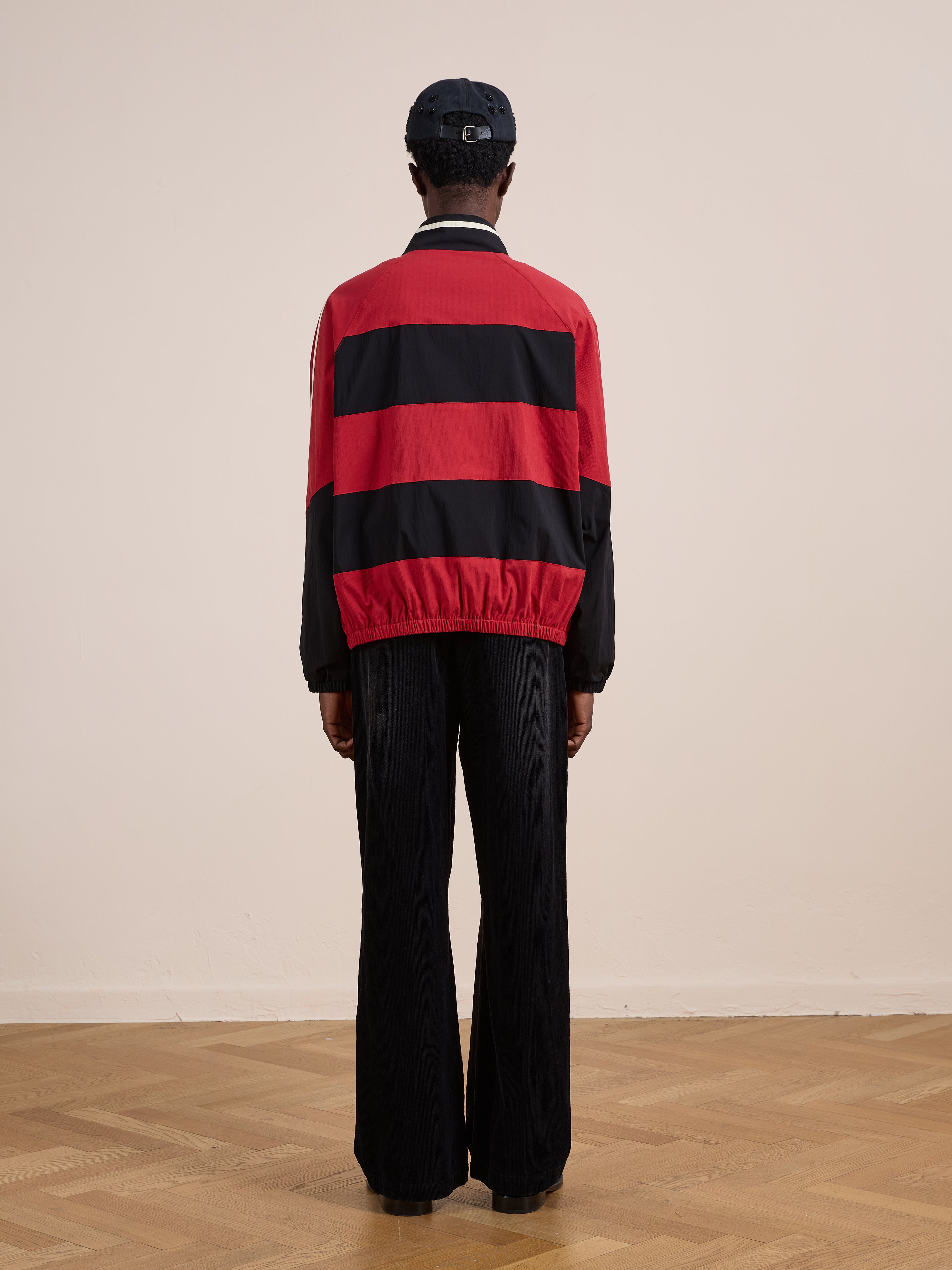 A person stands indoors facing away, wearing a black cap, black pants, and the FOUND Racing Striped Windbreaker in red and black on a wooden floor against a light-colored wall.