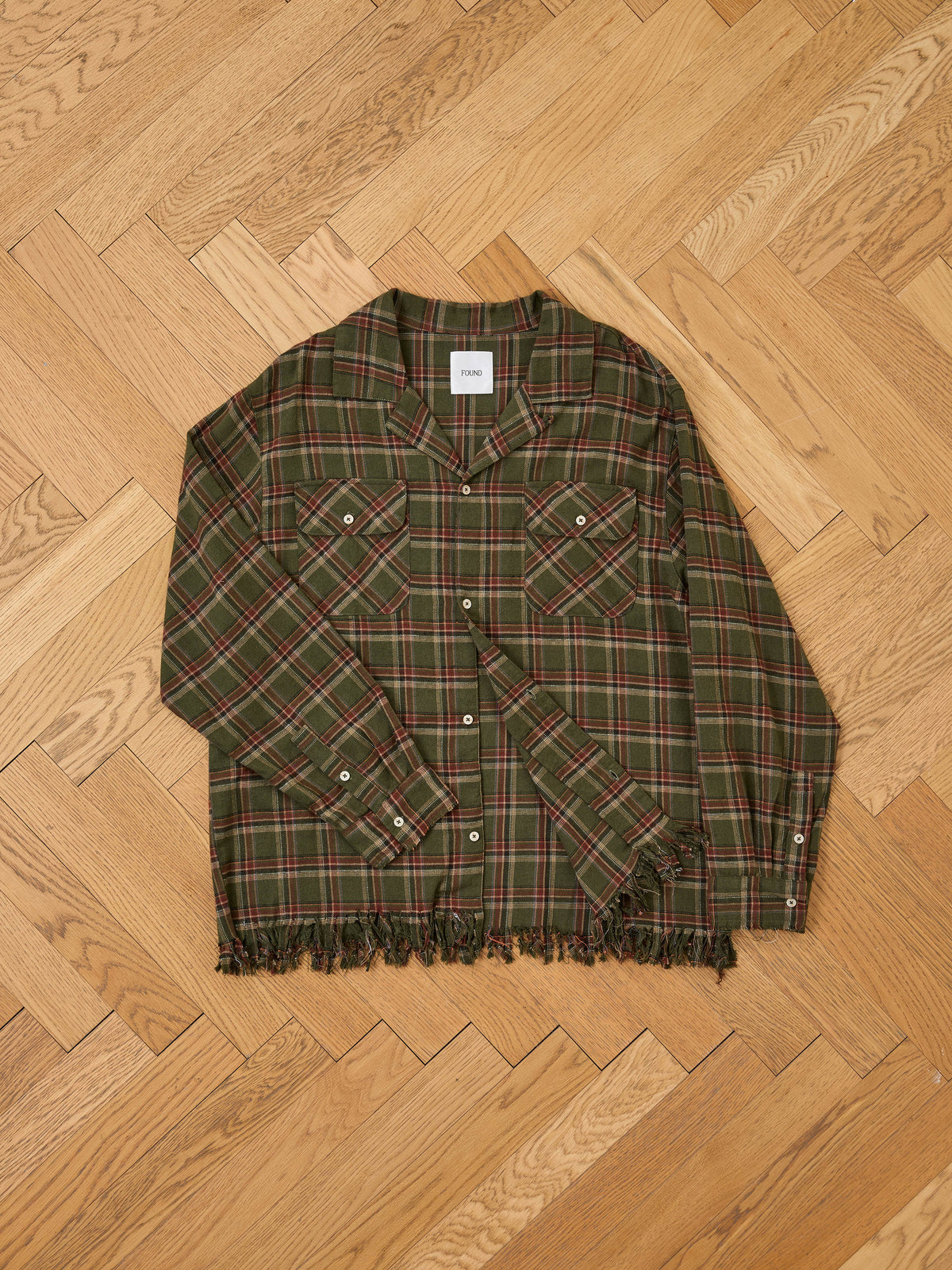 Raw Cut Fringe Plaid Camp Shirt by FOUND in green and red plaid with frayed hem, laid flat on a light herringbone-patterned wood floor.