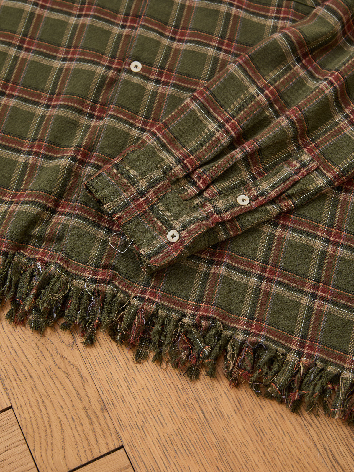 The FOUND Raw Cut Fringe Plaid Camp Shirt, featuring green and red plaid with white buttons and frayed edges, is displayed on a wooden surface.