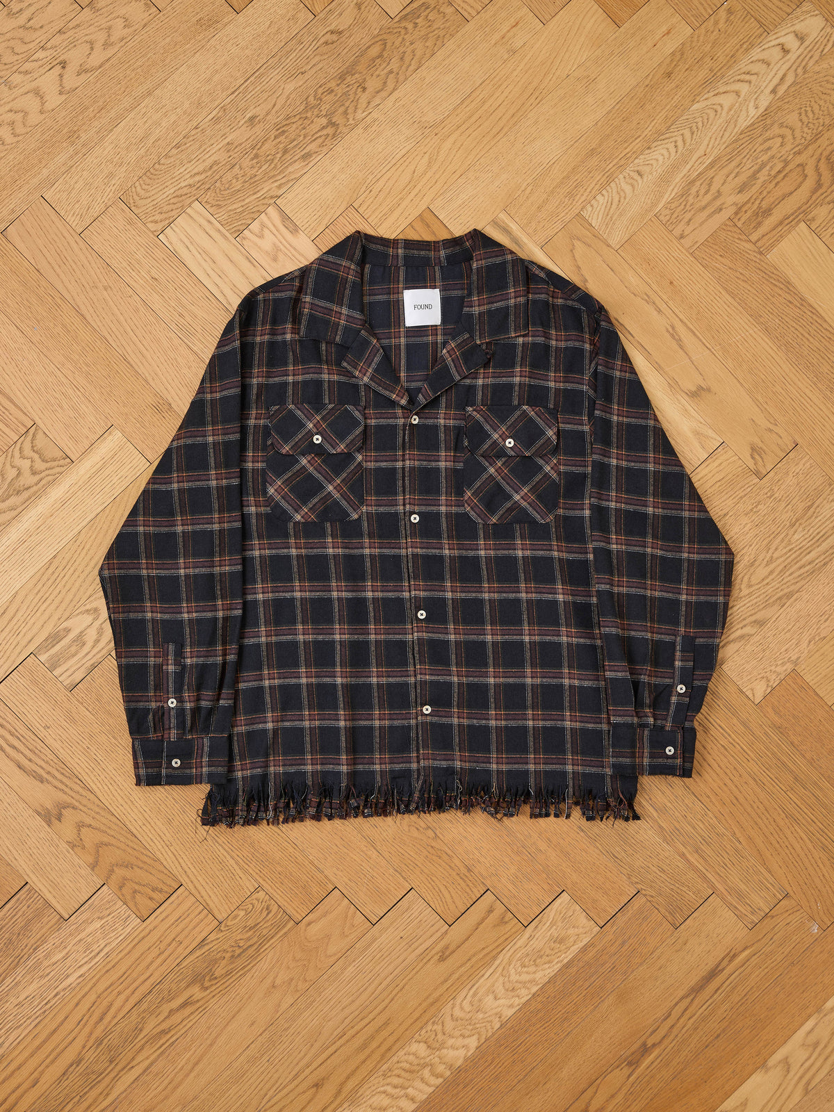 The FOUND Raw Cut Fringe Plaid Camp Shirt features long sleeves, dual chest pockets, button front, and a raw cut fringe hem, displayed on a wooden floor.