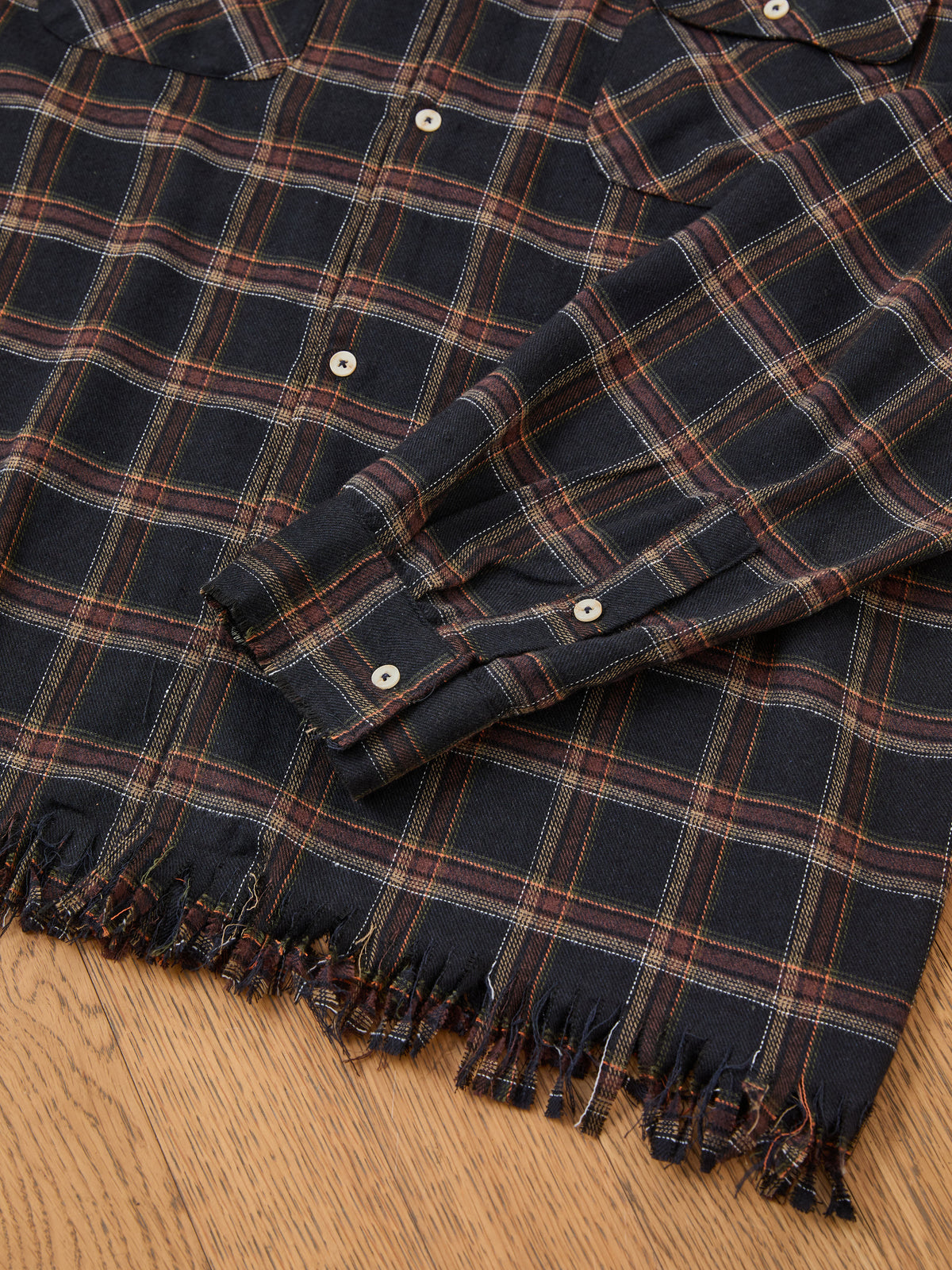 A close-up of the FOUND Raw Cut Fringe Plaid Camp Shirt with buttoned cuffs, featuring raw fringe, displayed flat on a wooden surface.