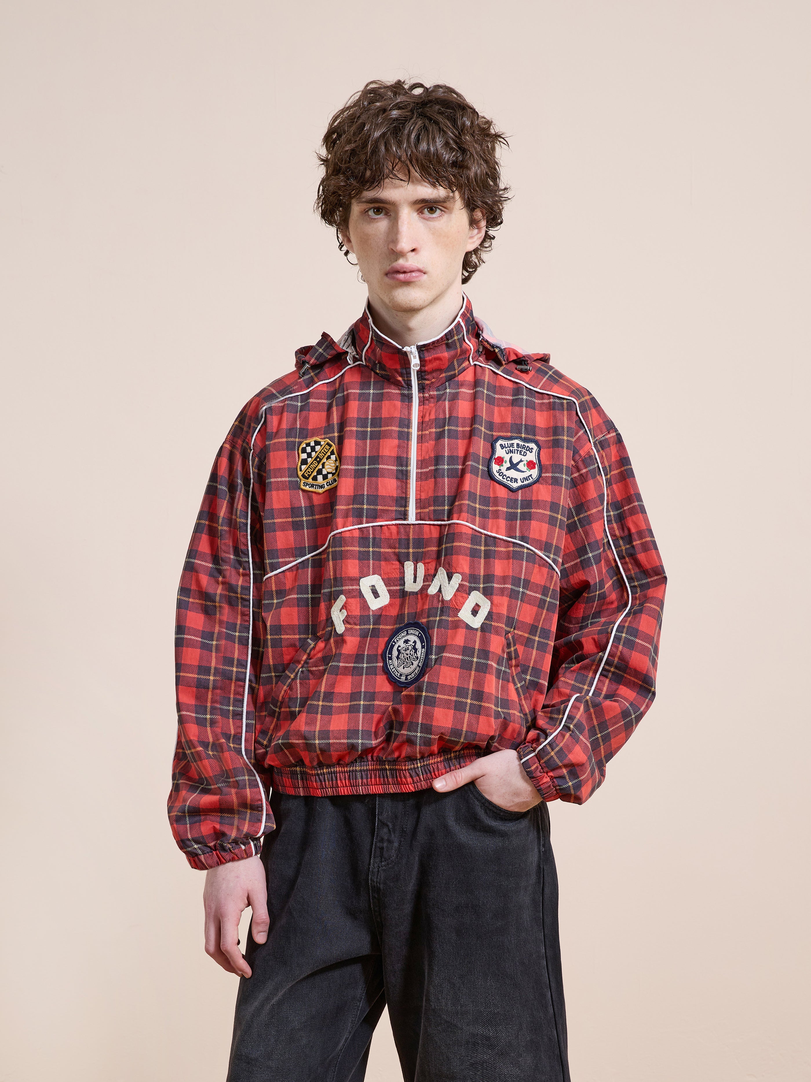 A young man with curly hair wears the FOUND Red Plaid Track Jacket, featuring a hood and patches, standing against a plain beige background.