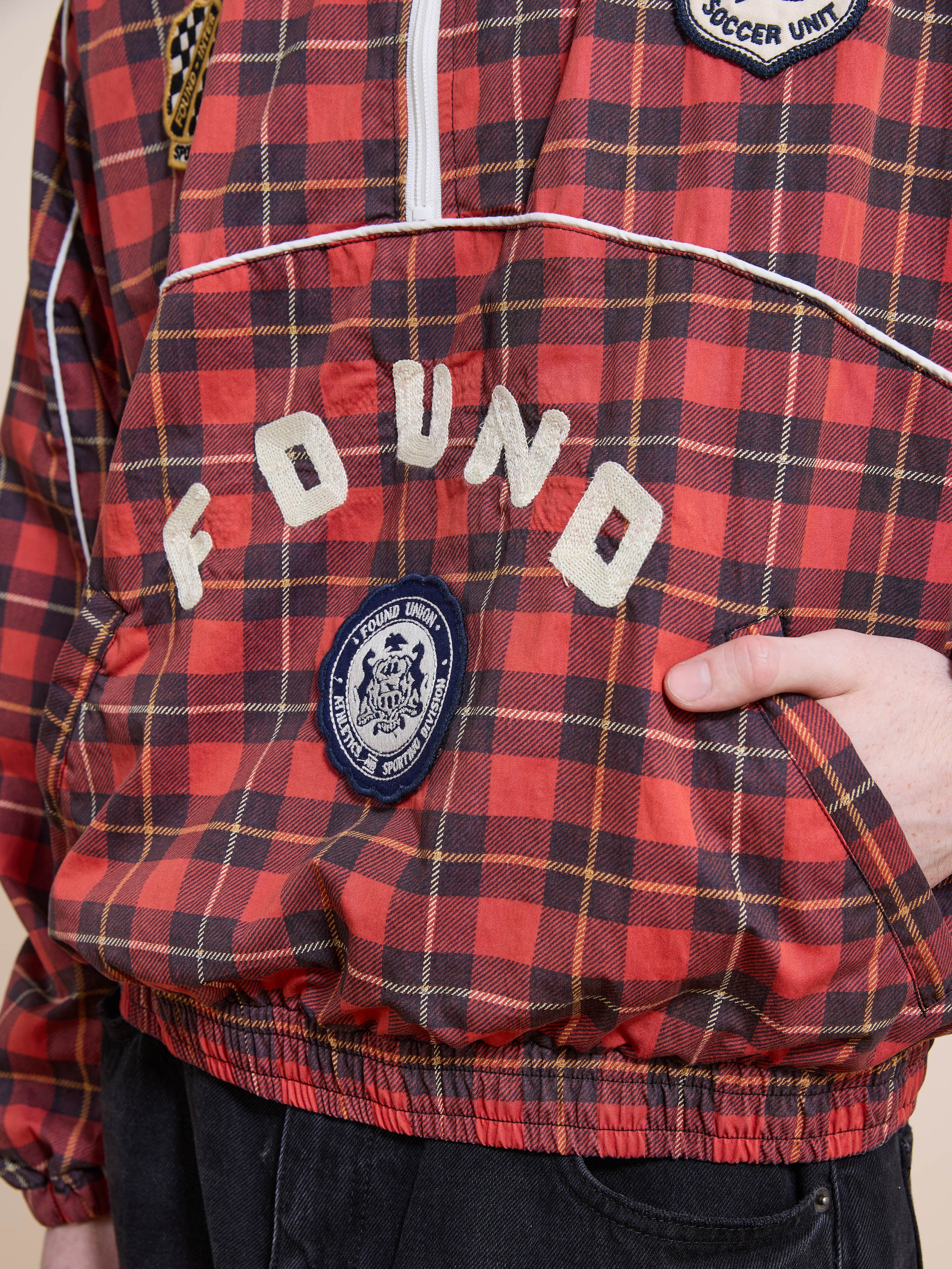 A person wears the FOUND Red Plaid Track Jacket with patches on the front and a hand in the pocket. Its unisex relaxed fit creates a laid-back vibe.