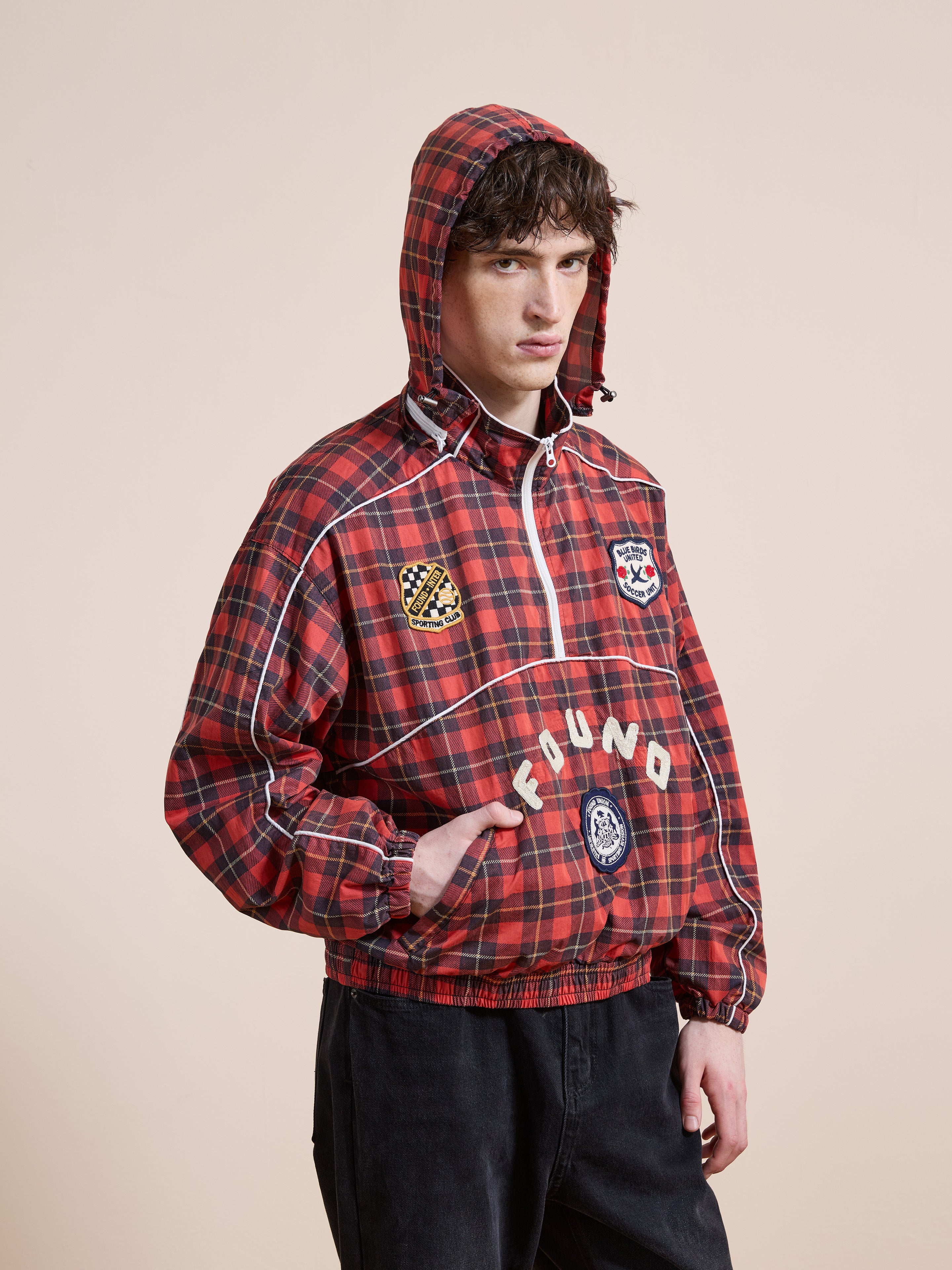Someone stands against a plain beige background wearing the FOUND Red Plaid Track Jacket, a unisex relaxed-fit piece featuring multiple patches and the brand name "FOUND" on the front.