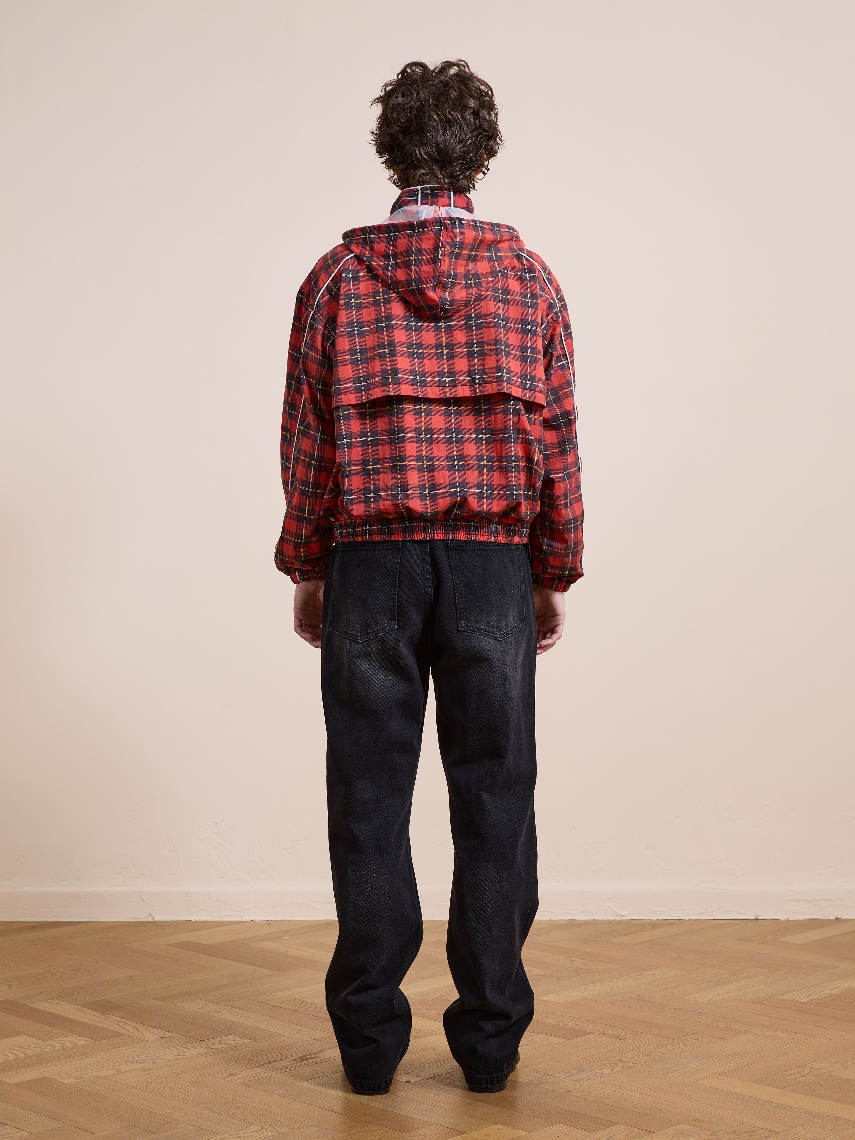 A person stands on a wooden floor indoors, facing a beige wall, wearing the FOUND Red Plaid Track Jacket styled like vintage sportswear and paired with black jeans.