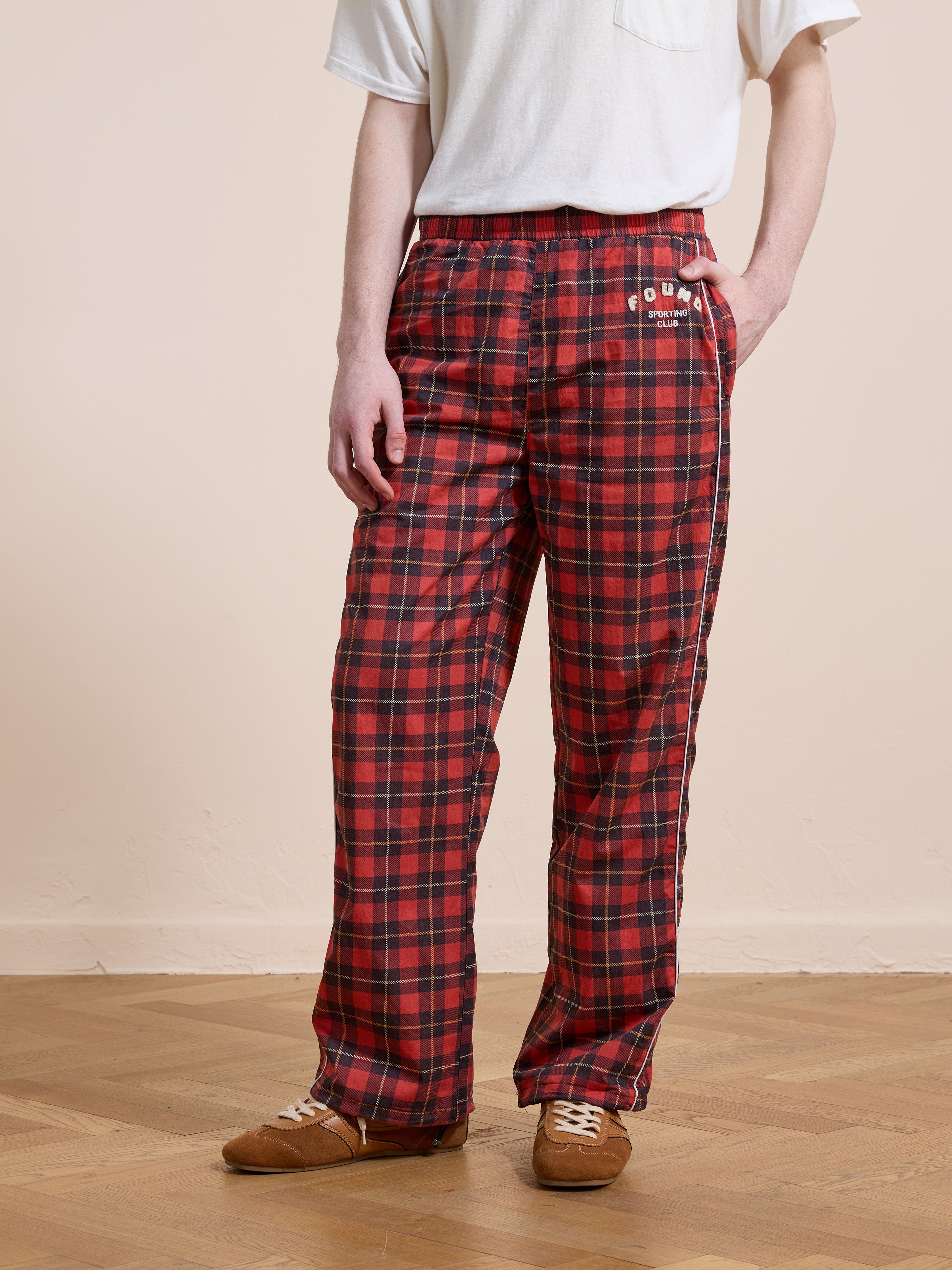 A person wears Found Red Plaid Track Pants in a unisex relaxed fit, featuring an embroidered logo, paired with a white t-shirt and brown shoes, standing on a wooden floor against a beige wall.