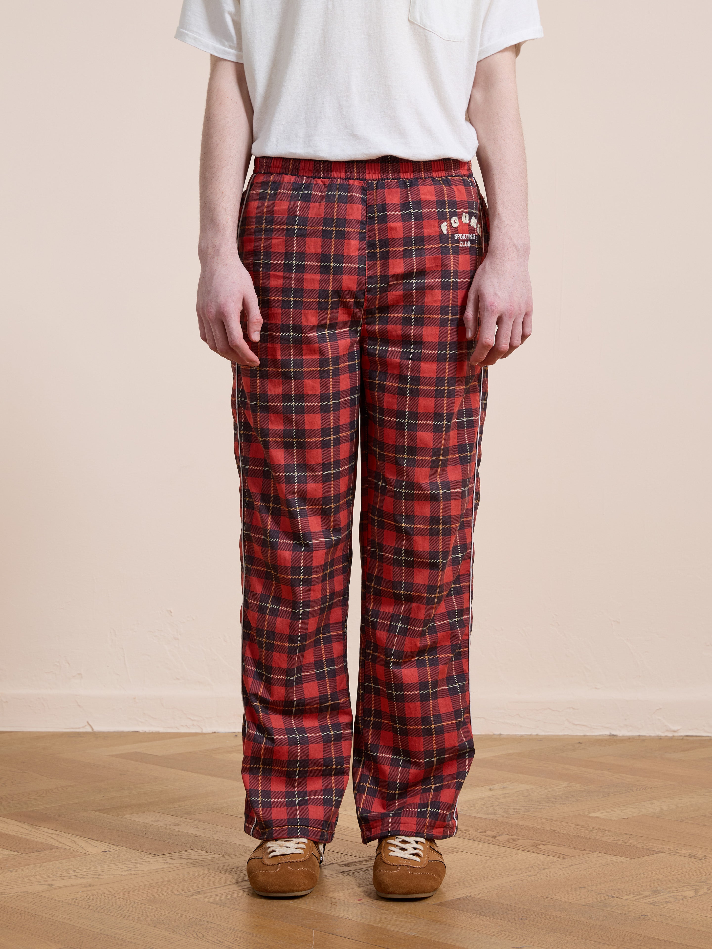 A person wearing Found Red Plaid Track Pants and a white short-sleeve t-shirt stands on a wooden floor against a plain beige wall.