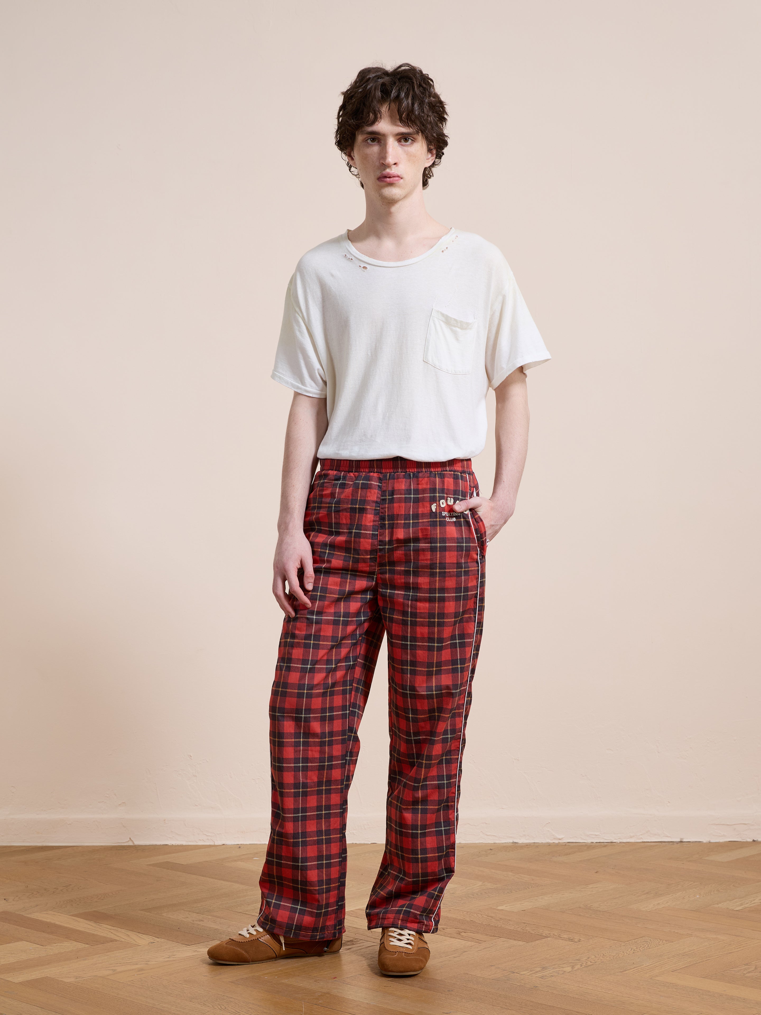 A person stands indoors against a beige wall and wooden floor, wearing Found's Red Plaid Track Pants with a relaxed fit, paired with a white pocket t-shirt and brown shoes.
