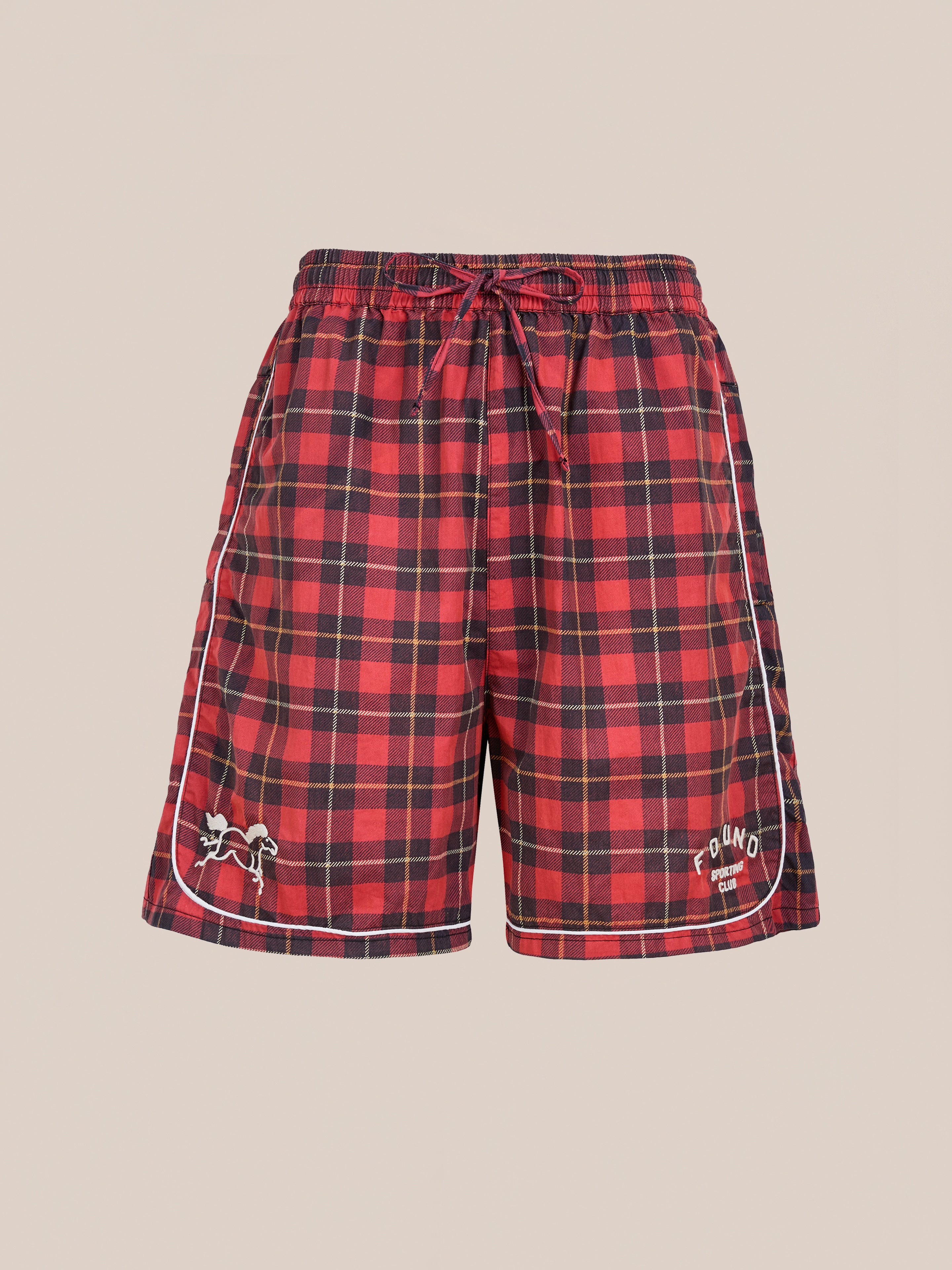 Found Red Plaid Track Shorts feature black plaid, white piping, and embroidered details on the left hem and right thigh. These vintage-inspired unisex shorts offer a relaxed fit and are shown on a plain beige background.