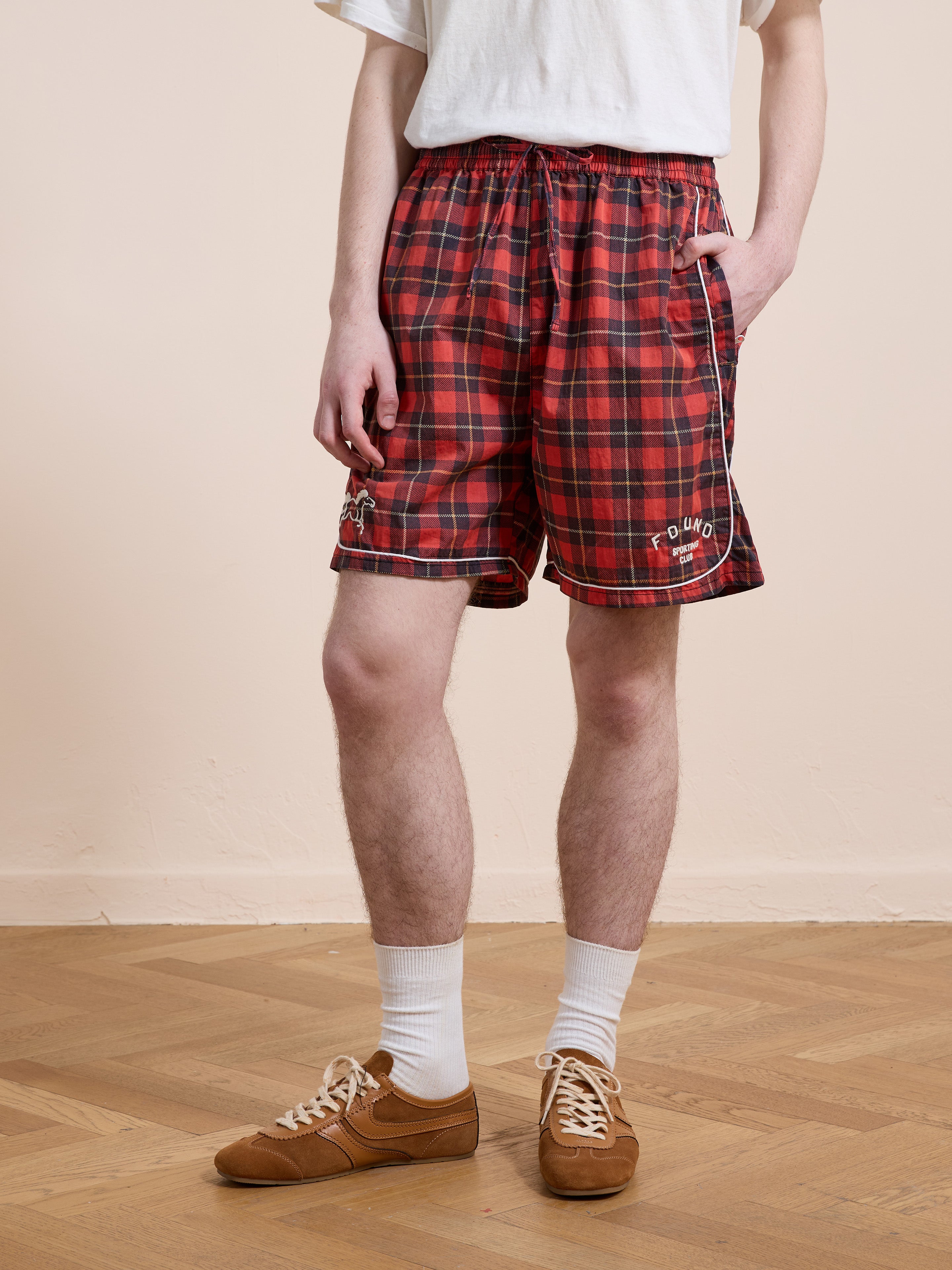 A person wearing a white t-shirt, Found Red Plaid Track Shorts, white socks, and brown sneakers stands on a wooden floor with one hand in their pocket.