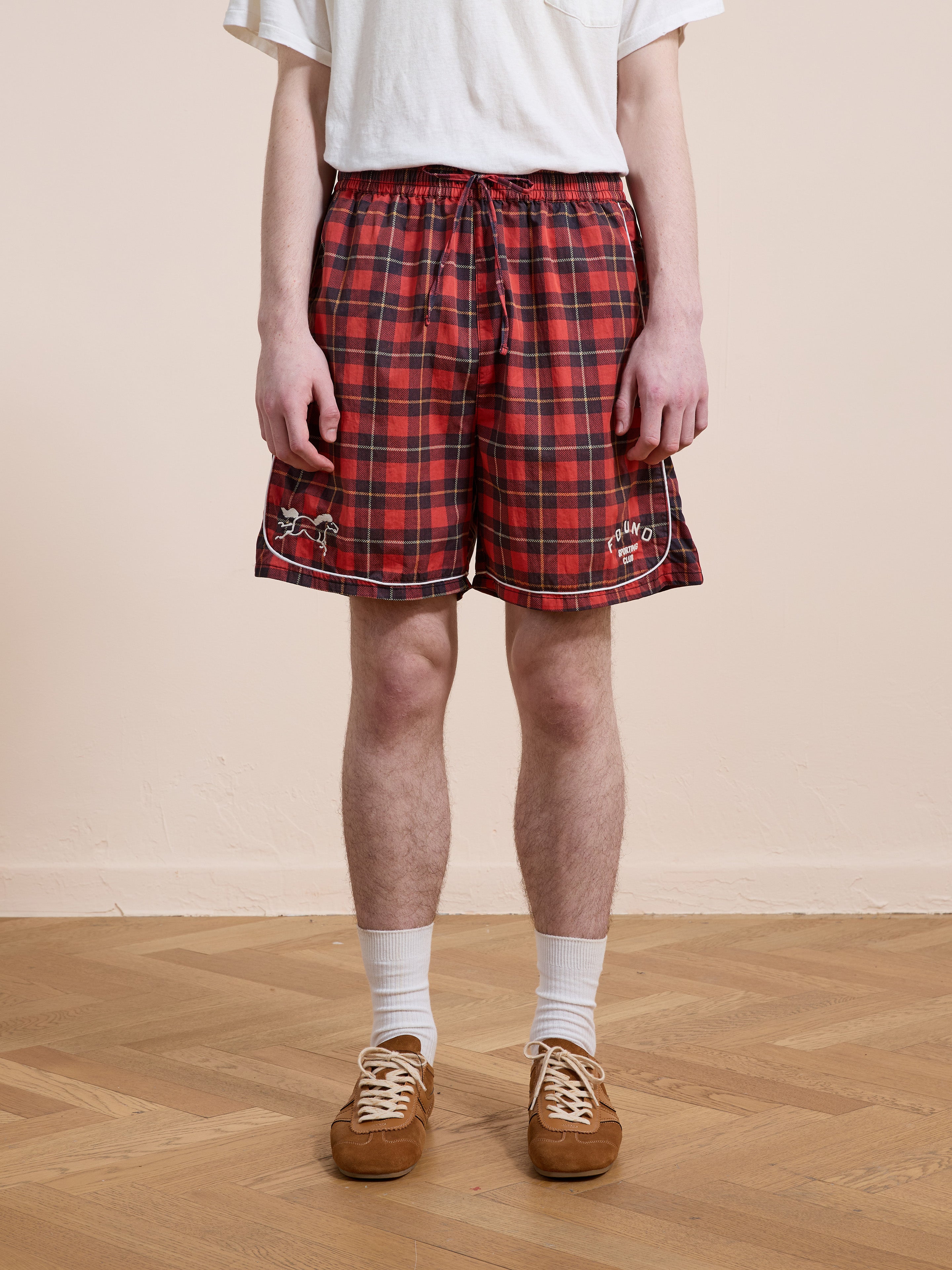 Indoors, a person wears a white t-shirt, Found Red Plaid Track Shorts (unisex relaxed fit with embroidery), white socks, and brown sneakers on a wooden floor.