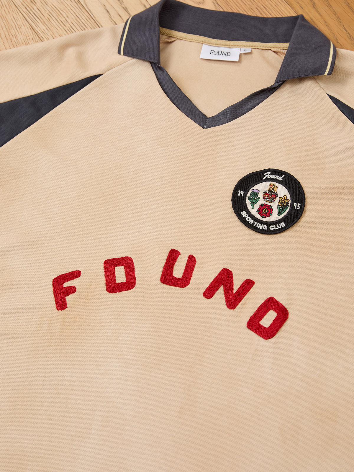 A standout piece of unisex sportswear, the Rosewood Jersey by FOUND features a beige vintage football design with black collar and sleeves, “FOUND” in bold red letters, and a circular “Found Sporting Club” patch on the chest—laid flat on a wooden surface.