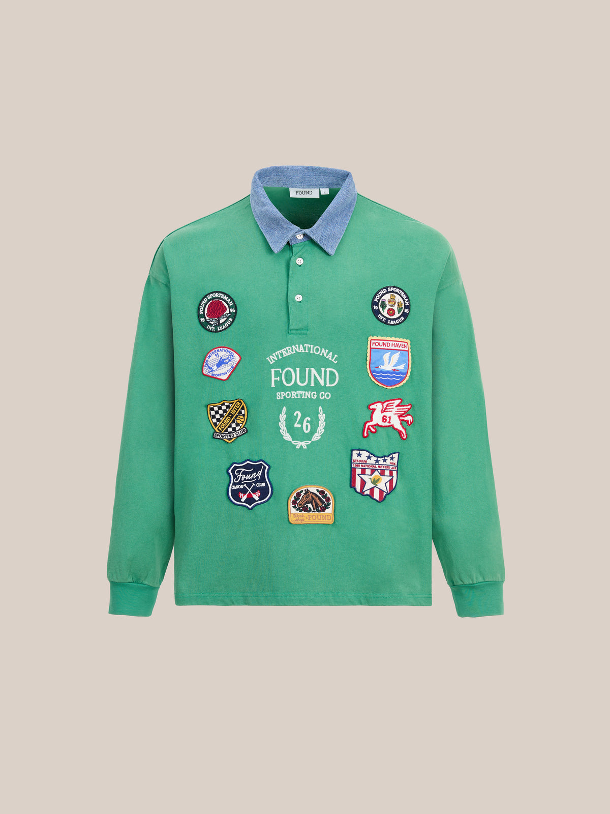 The FOUND Rugby Polo Shirt is a green long-sleeve crafted from midweight cotton, designed with a blue denim collar, vibrant embroidered patches, and bold white “INTERNATIONAL FOUND SPORTING CO” lettering across the front for a classic collegiate sport-inspired look.