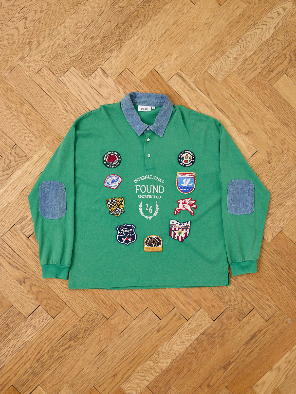 FOUND Rugby Polo Shirt in green, featuring long sleeves and crafted from midweight cotton with a denim collar and elbow patches. Reminiscent of a classic rugby shirt, it’s adorned with multiple colorful embroidered patches on the front and is shown laid flat on a wooden herringbone floor.