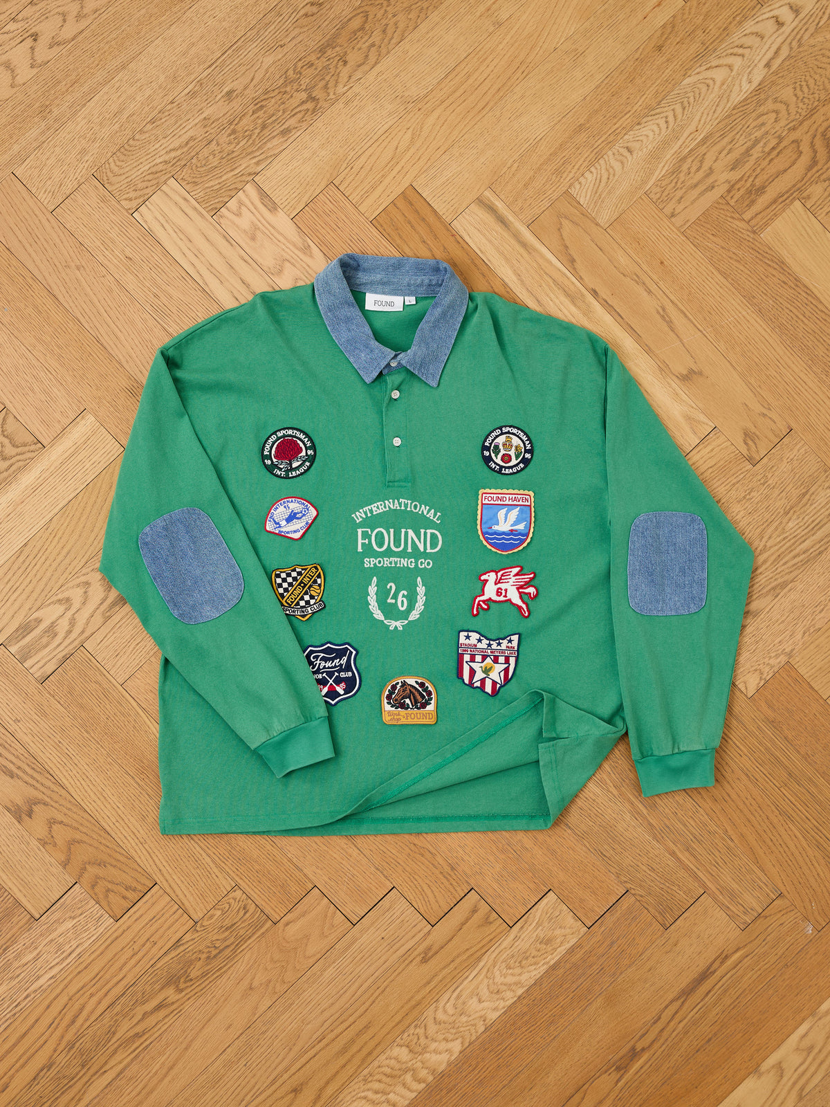 The FOUND Rugby Polo Shirt, featuring green long sleeves, a blue denim collar, and contrasting elbow patches, is crafted from midweight cotton and adorned with an array of vibrant embroidered patches on the front. It is shown laid flat on a wooden floor.