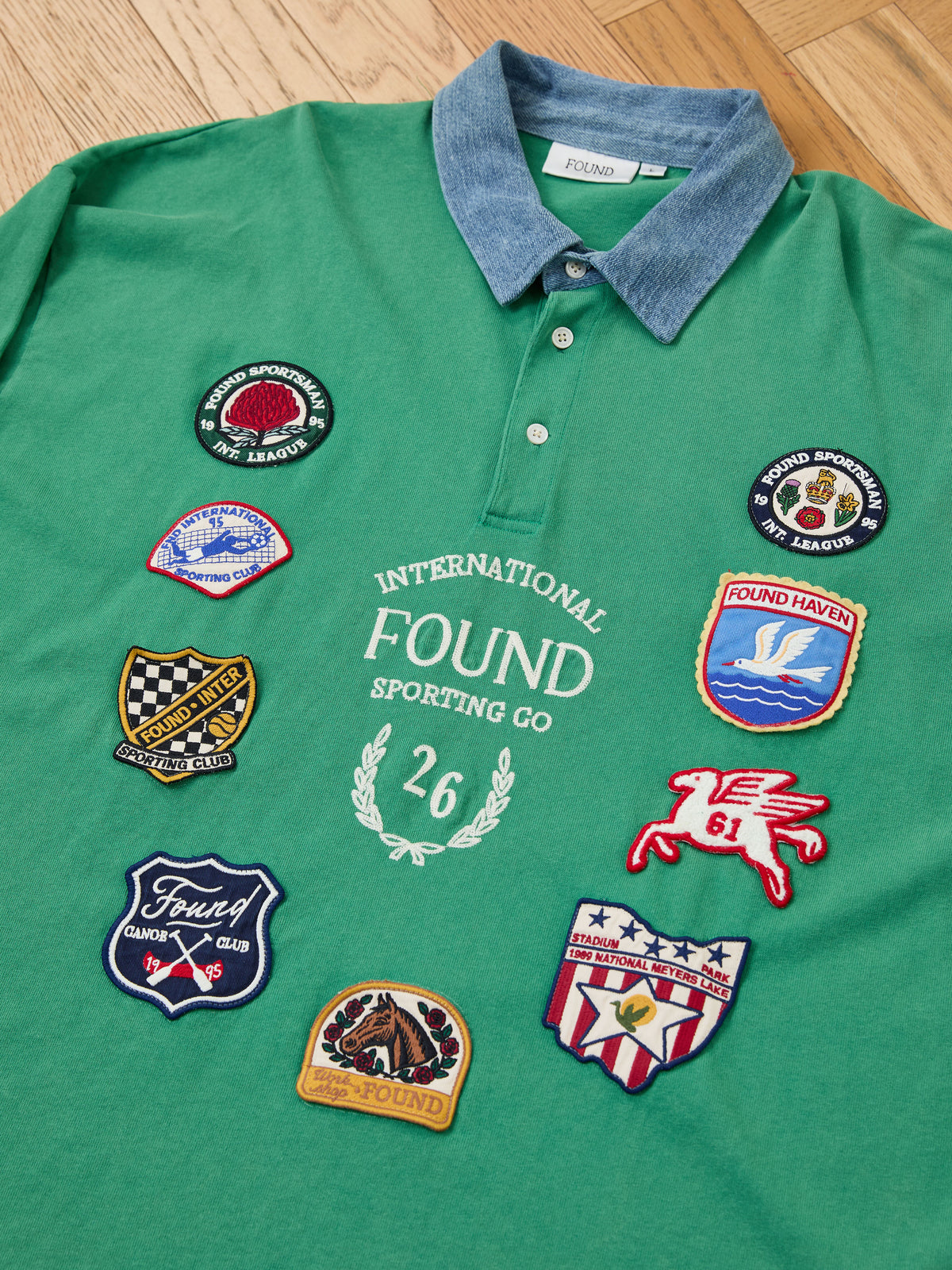 Introducing the FOUND Rugby Polo Shirt: a green midweight cotton polo featuring a classic collar, vibrant embroidered patches, and the phrase “International Found Sporting Co 26” on the front—capturing timeless collegiate sport style.